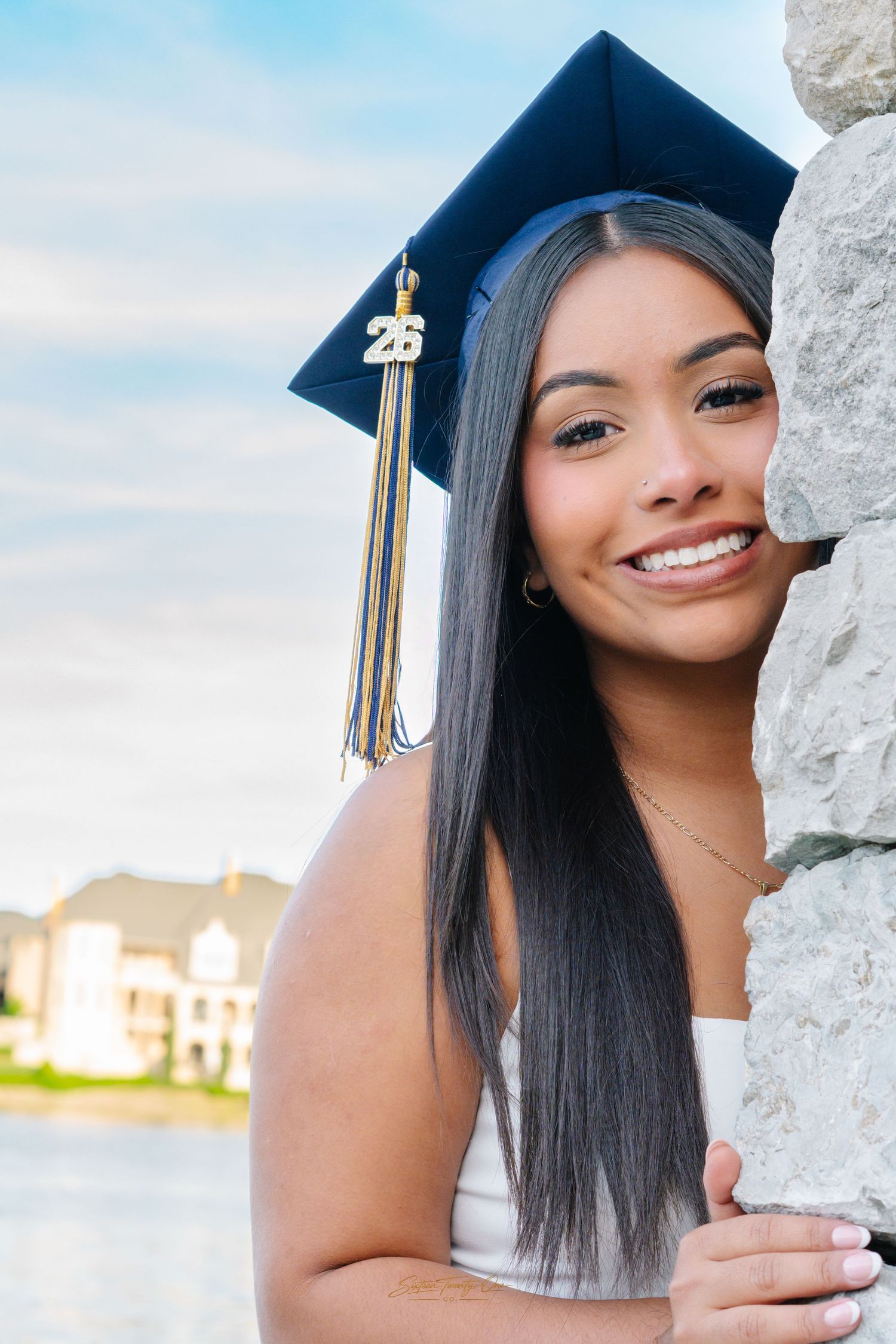 Smiling graduate in blue cap with 25 tassel leaning against a stone wall outdoors.