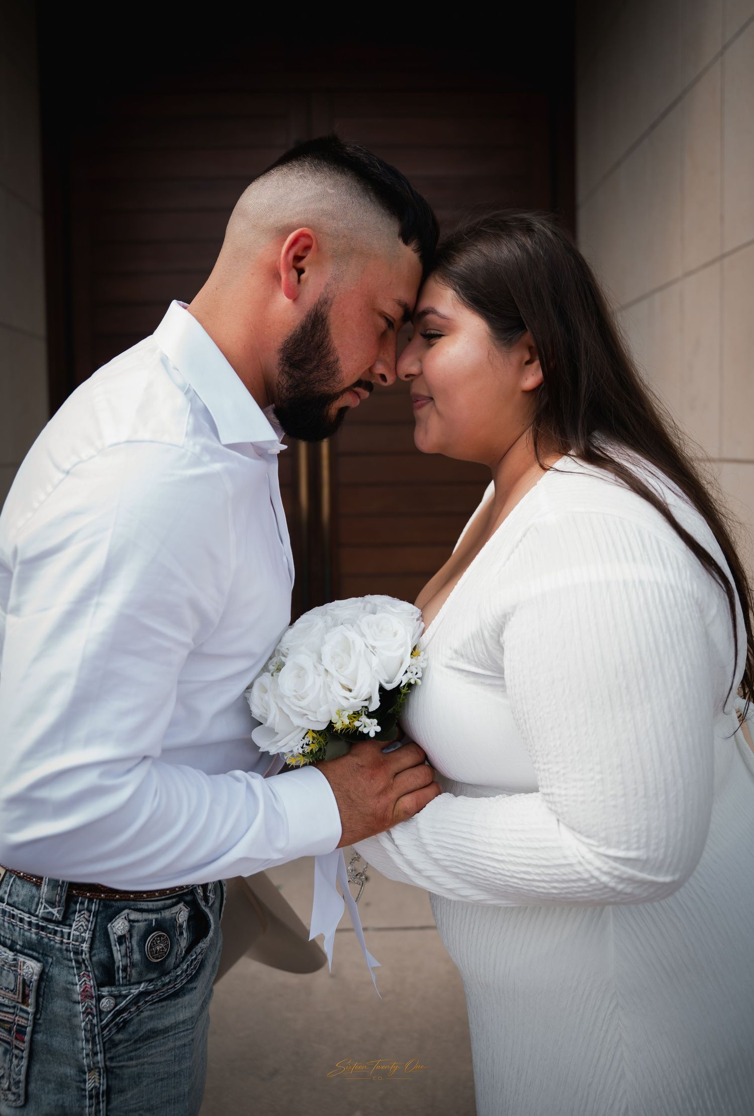 Couple in white wedding attire sharing an intimate moment with bride holding white rose bouquet.