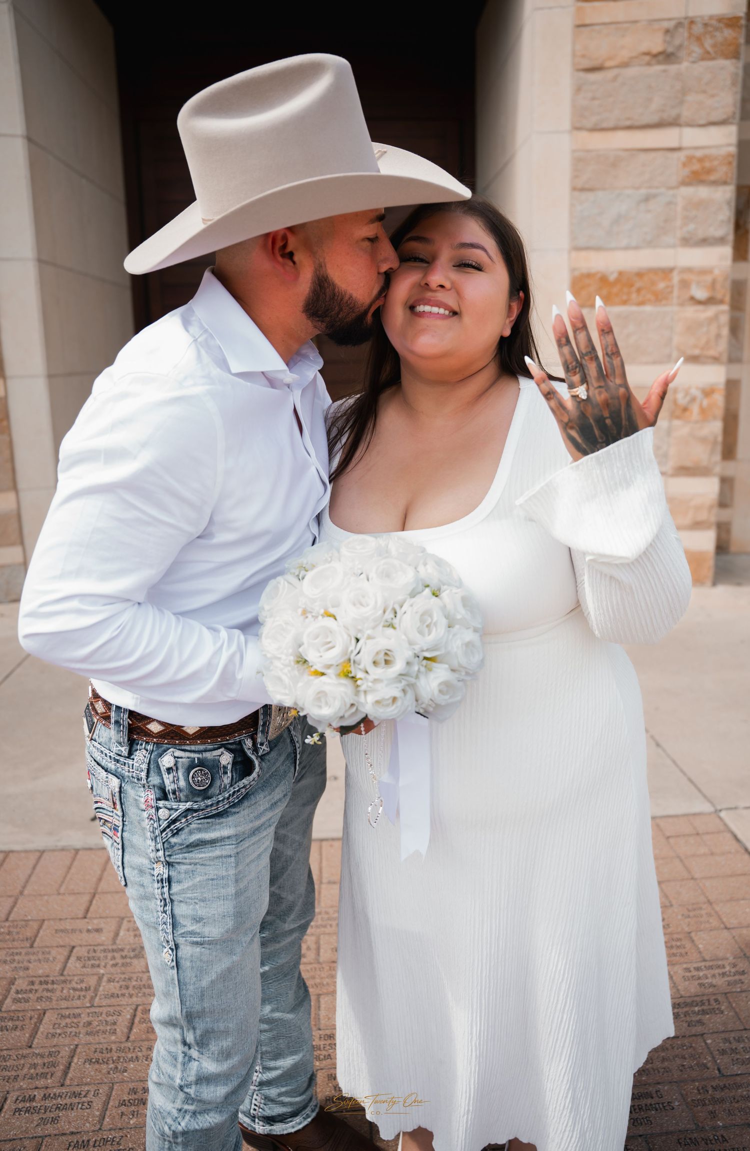 Groom in cowboy hat kissing bride's cheek as she shows off wedding ring, holding white rose bouquet.