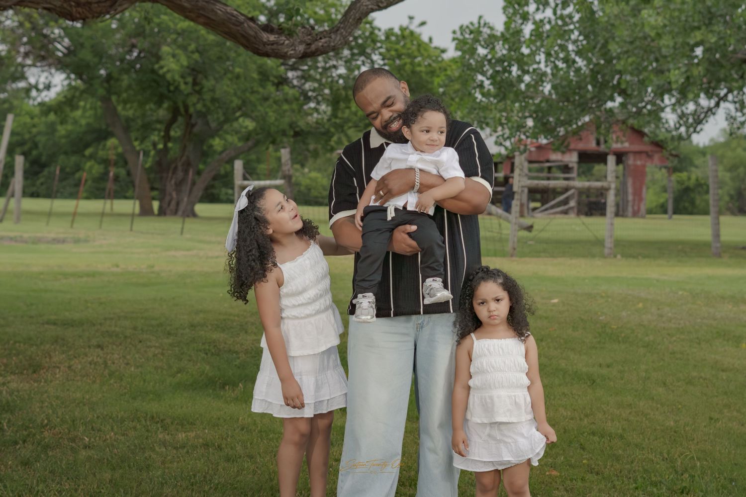 Father holding baby surrounded by two young daughters in white outfits outdoors.
