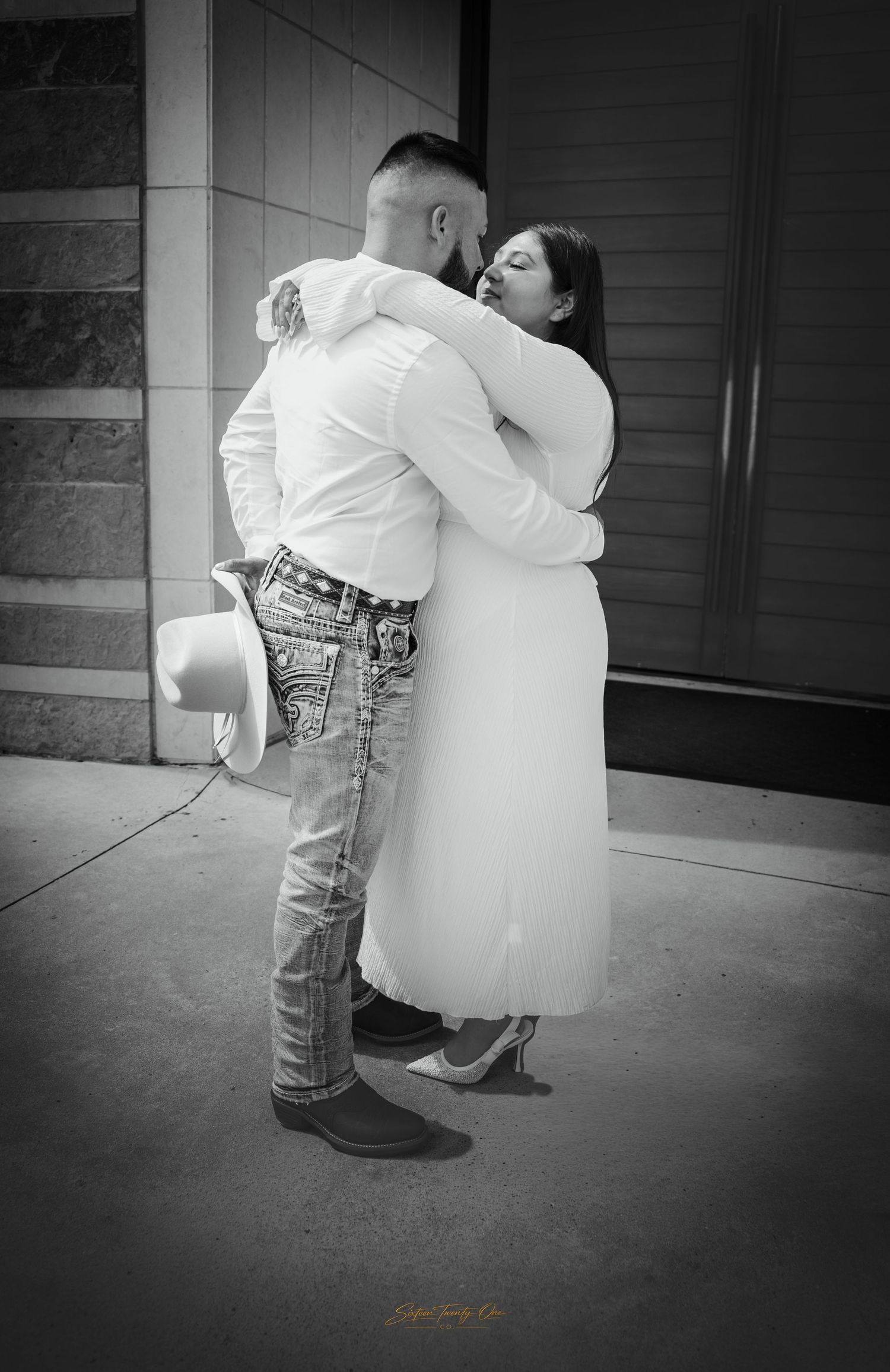 Black and white photo of newlyweds sharing passionate kiss outside, groom holding cowboy hat.