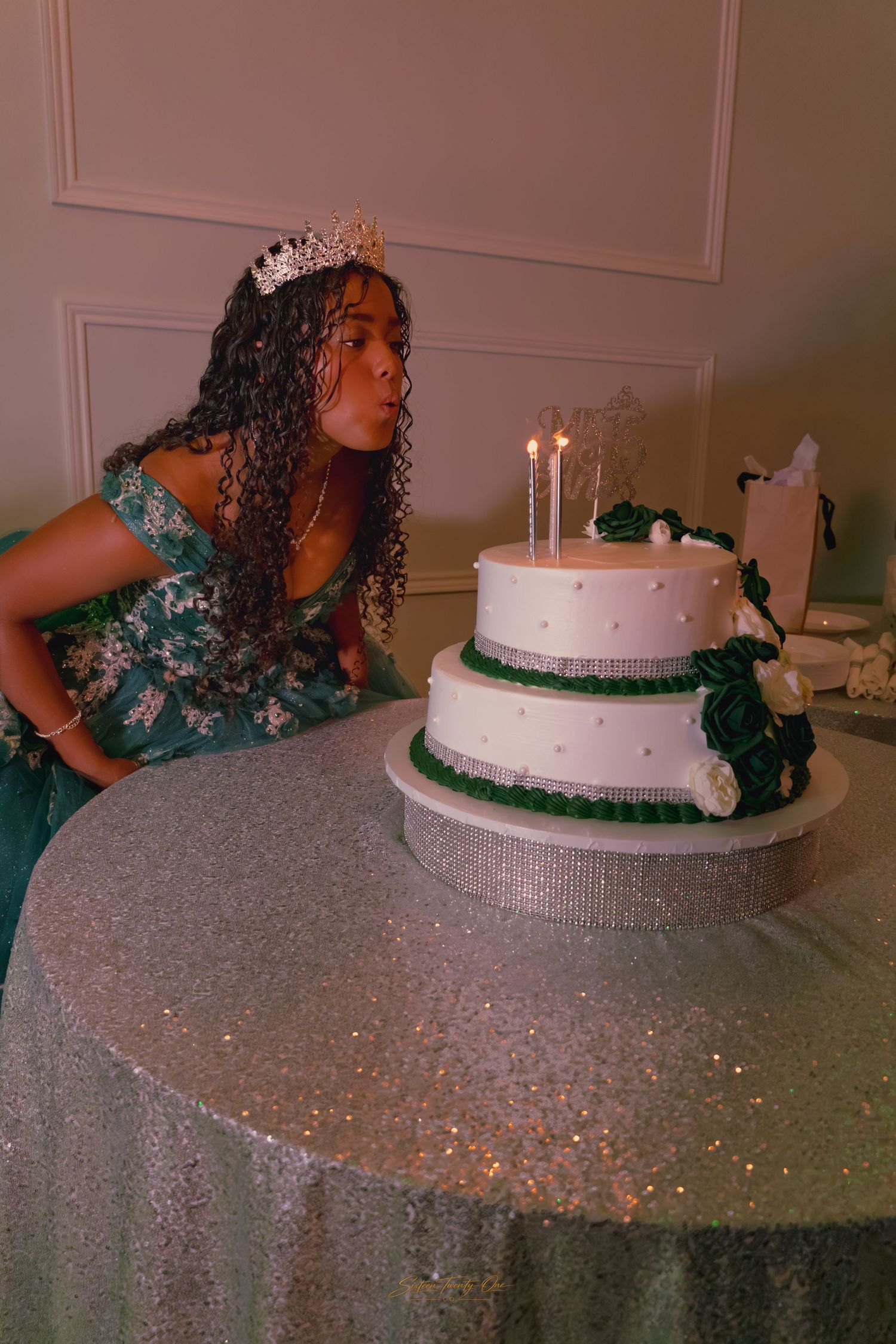 Young woman in green quinceañera gown and tiara blows out candles on a two-tier white cake.