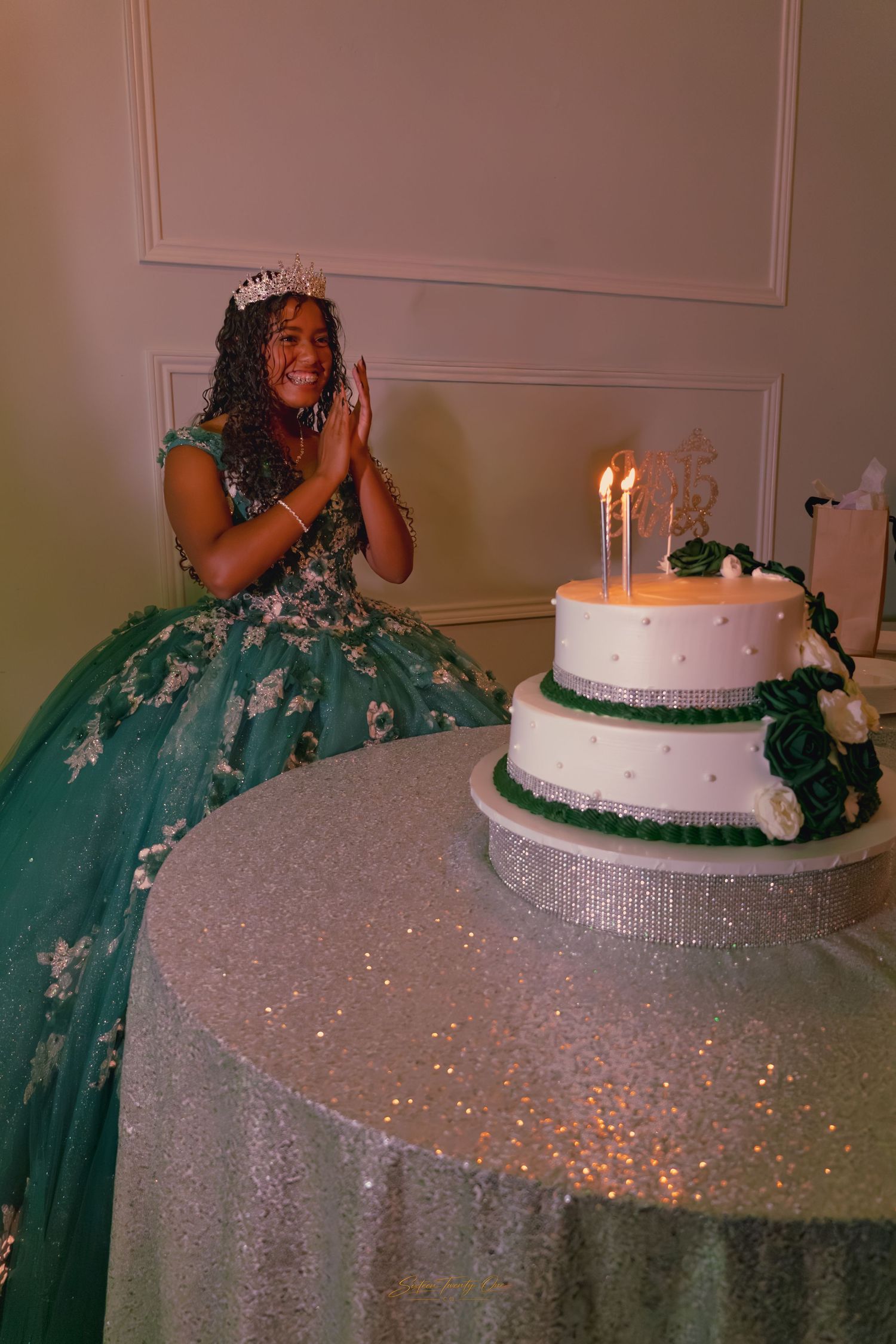 Girl in green quinceañera dress and crown claps excitedly beside a white two-tier birthday cake.