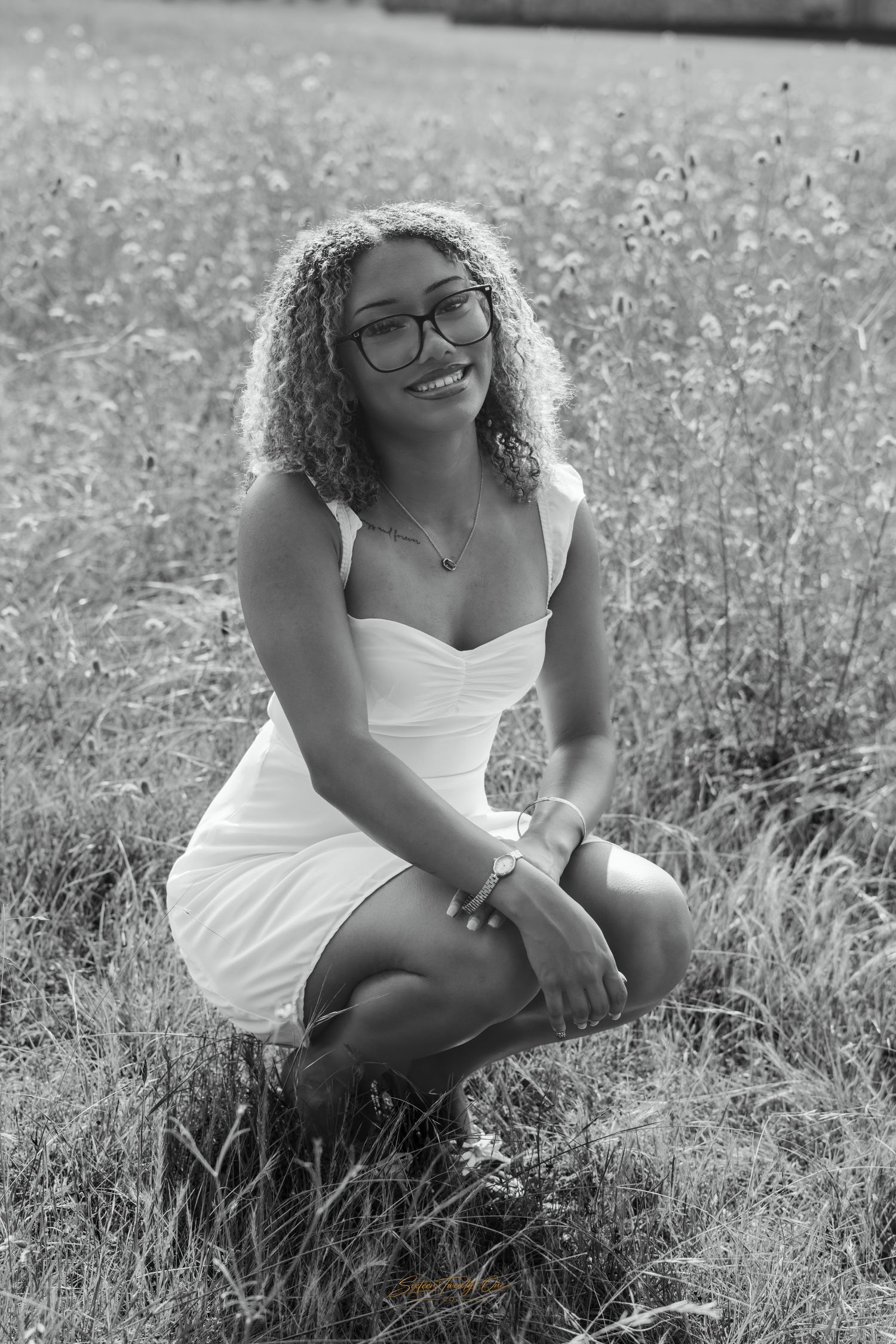 Black and white portrait of a smiling young woman in white dress crouching in a wildflower field.