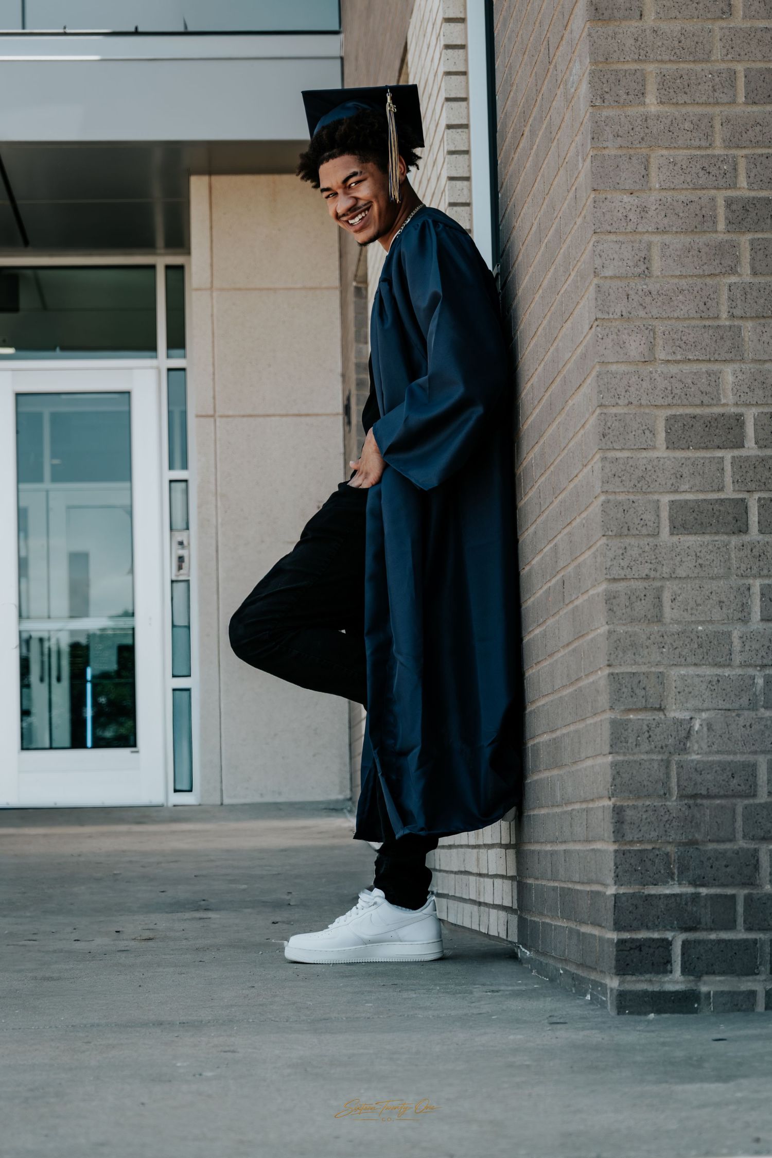 Smiling young man in navy graduation cap and gown leaning against brick wall outside school building.