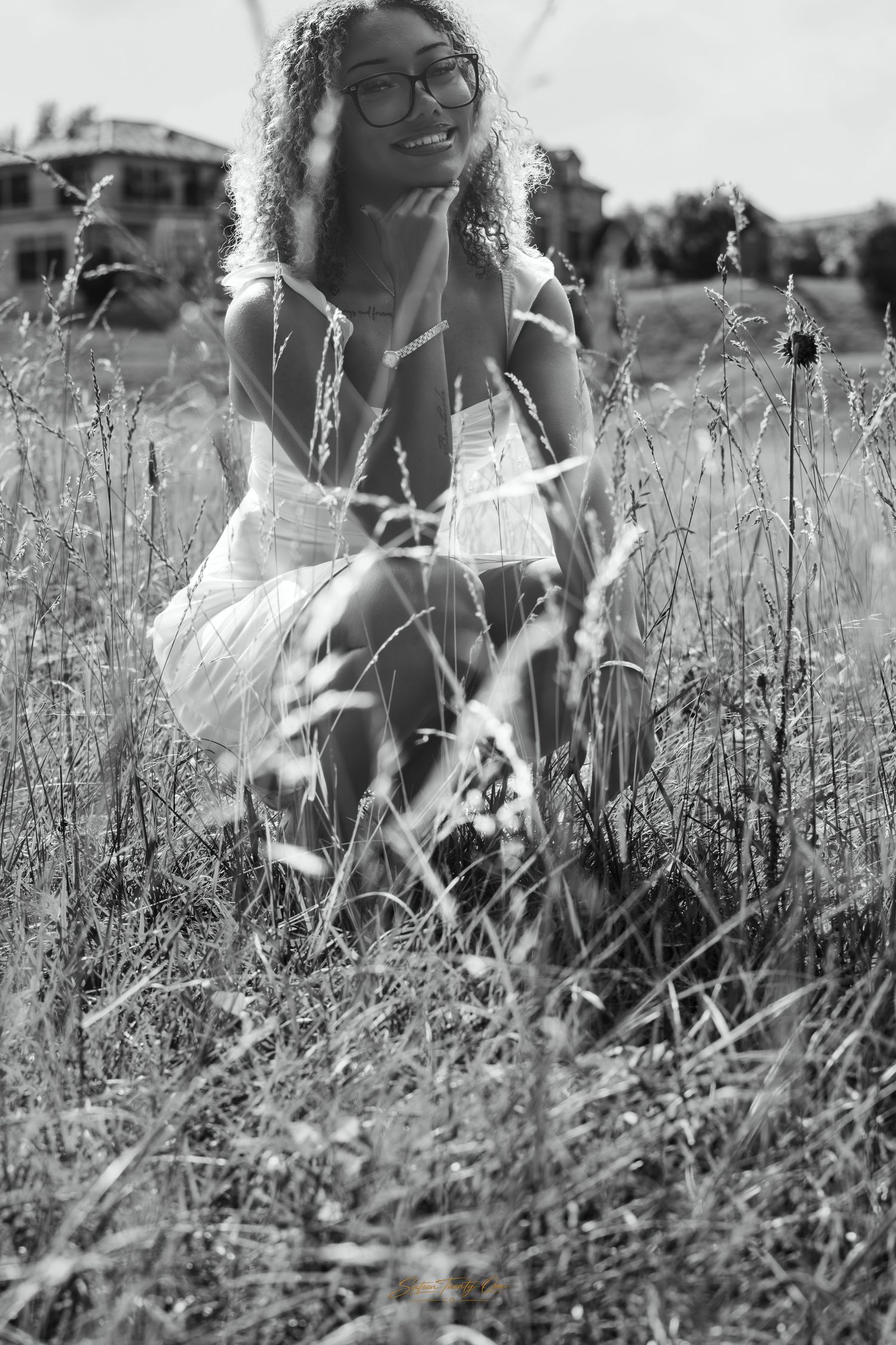 Black and white portrait of curly-haired woman in white dress crouching in tall grass.