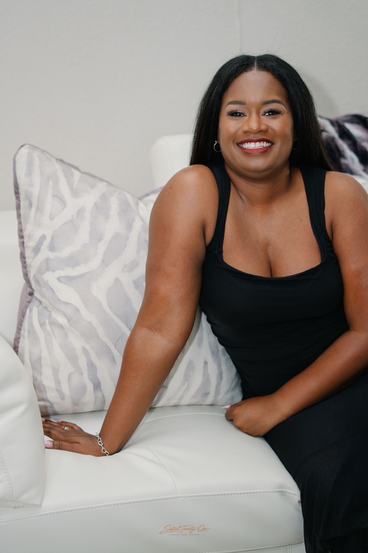 Smiling Black woman in black dress sitting on white sofa with decorative pillows.