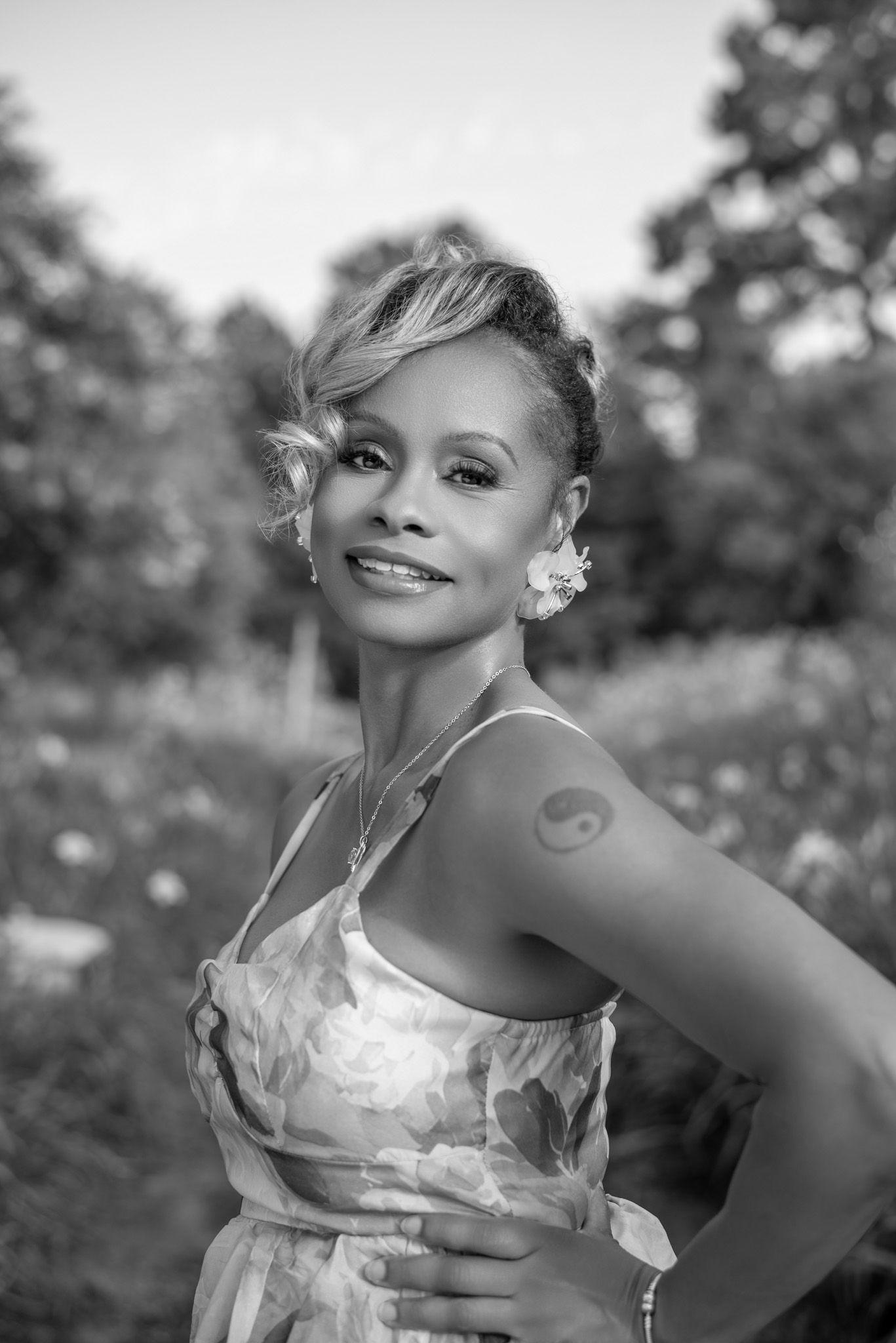 Black and white portrait of smiling woman with yin yang tattoo in floral dress outdoors.
