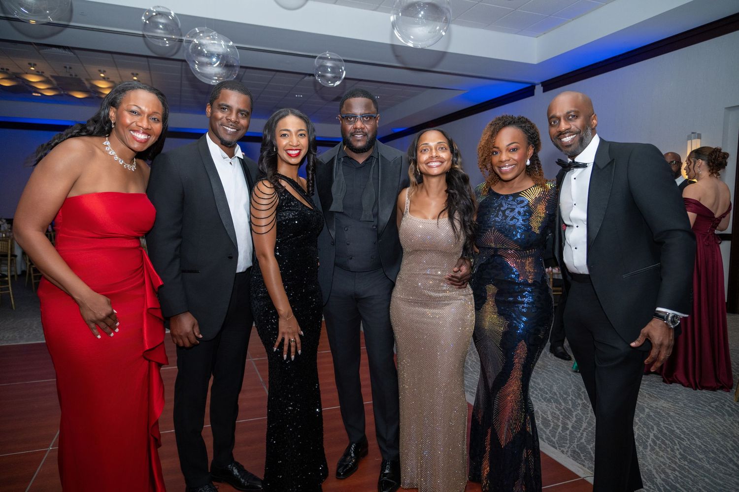 Group of seven elegantly dressed people posing together at a formal gala event in a banquet hall.