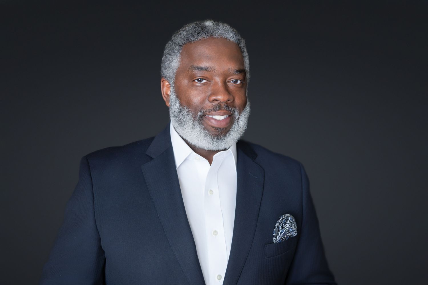 Professional portrait of a distinguished Black man with gray beard wearing a navy blazer and white shirt on dark background.