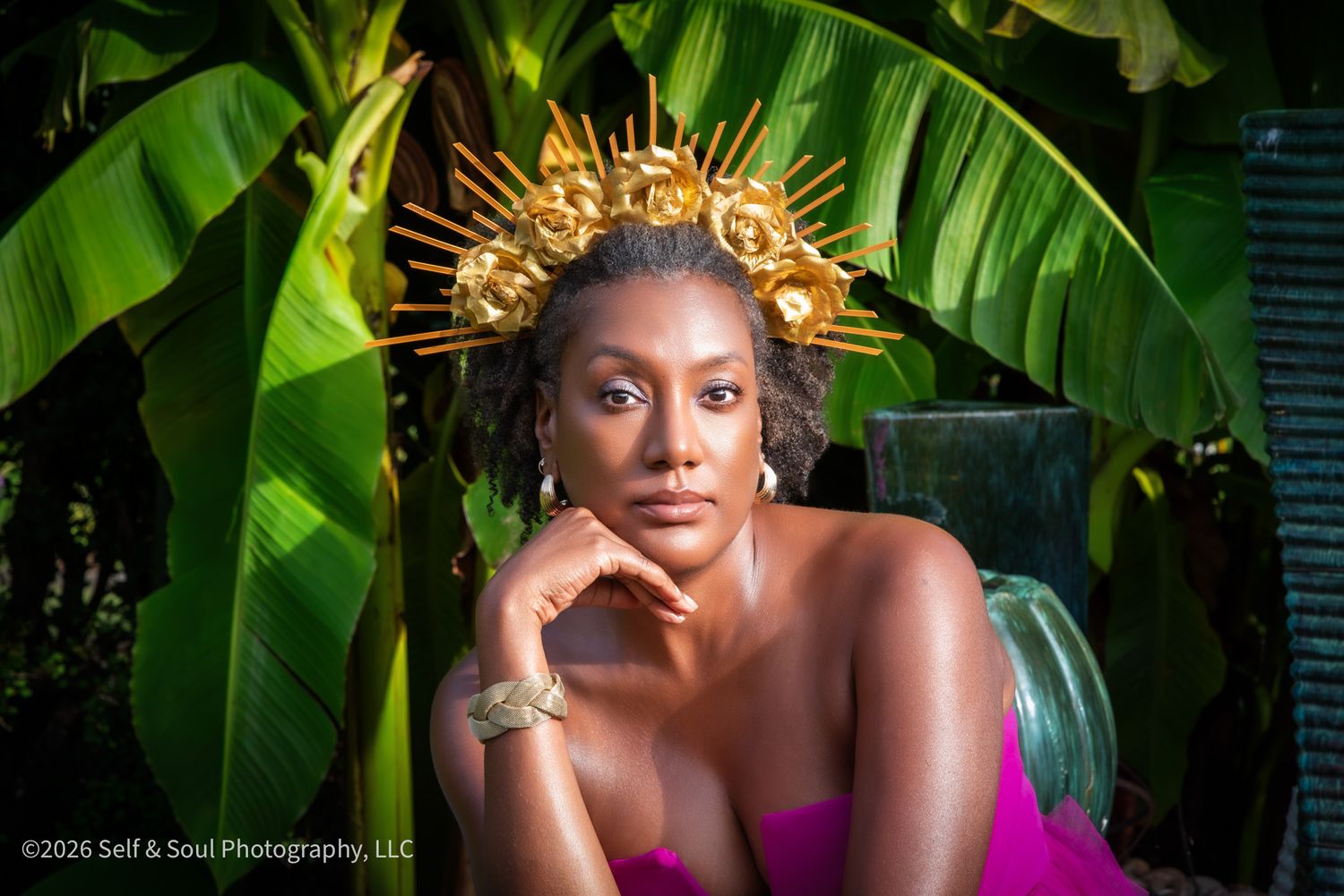 Woman wearing gold sunburst crown poses elegantly before tropical banana leaves in vibrant pink dress.
