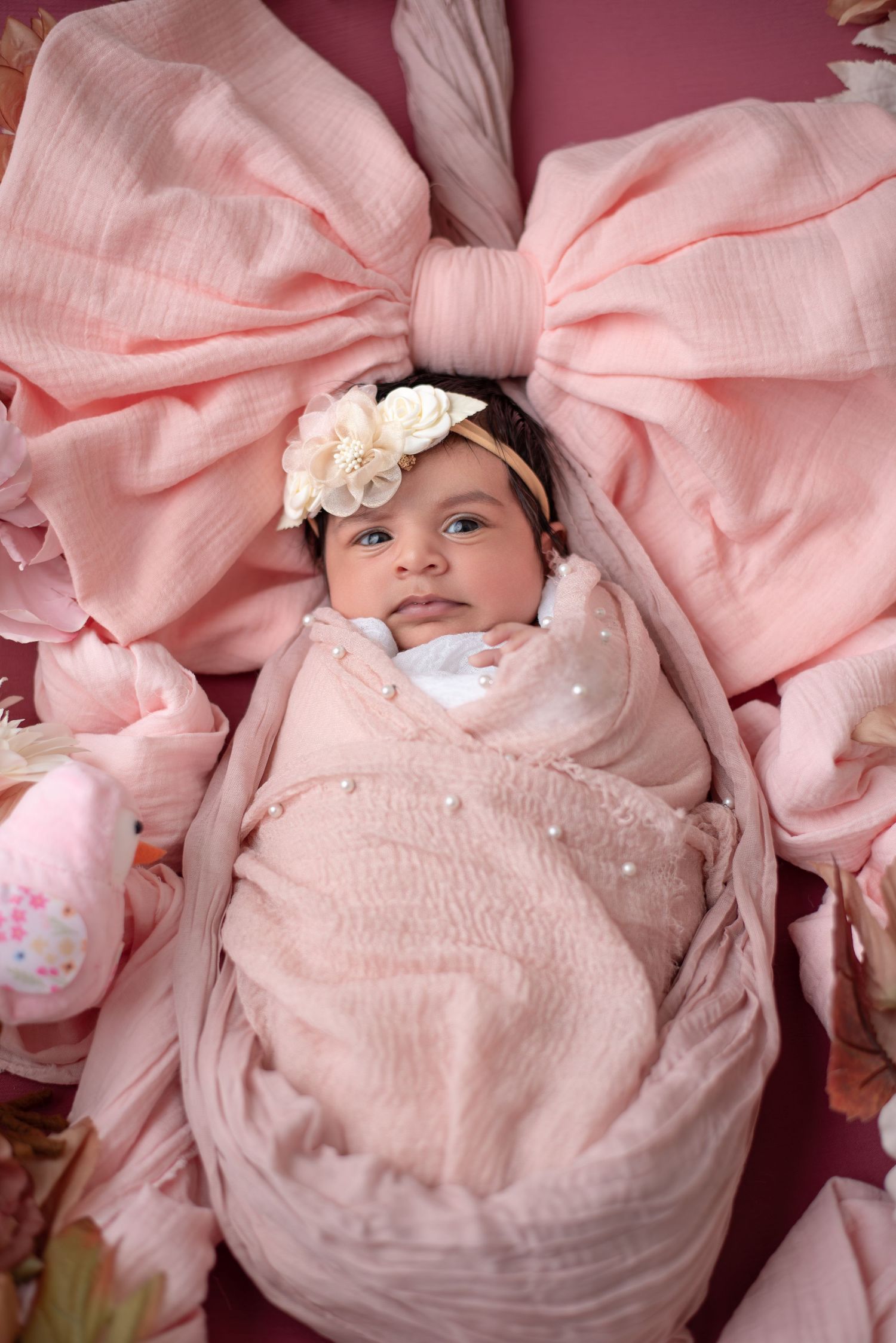 Newborn baby girl swaddled in pink muslin wrap with floral headband, posed on pink bow backdrop.