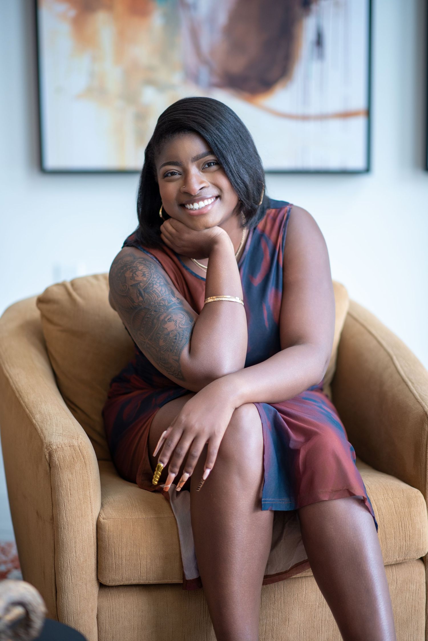 Smiling woman in colorful sleeveless dress sits in gold armchair, showing tattoo sleeve, gold jewelry, in bright modern room.
