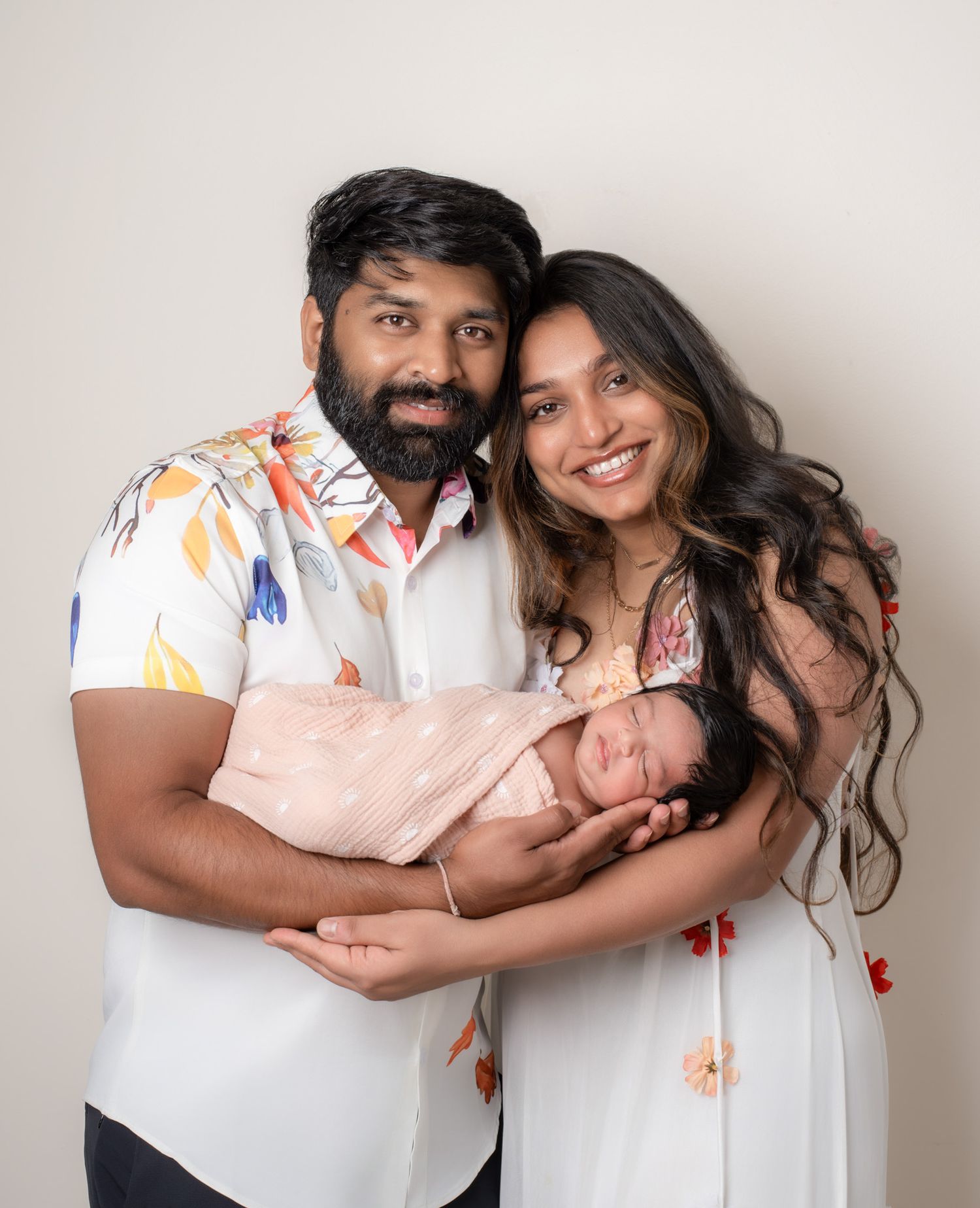 Happy couple smiling and holding their sleeping newborn baby wrapped in a pink swaddle blanket.