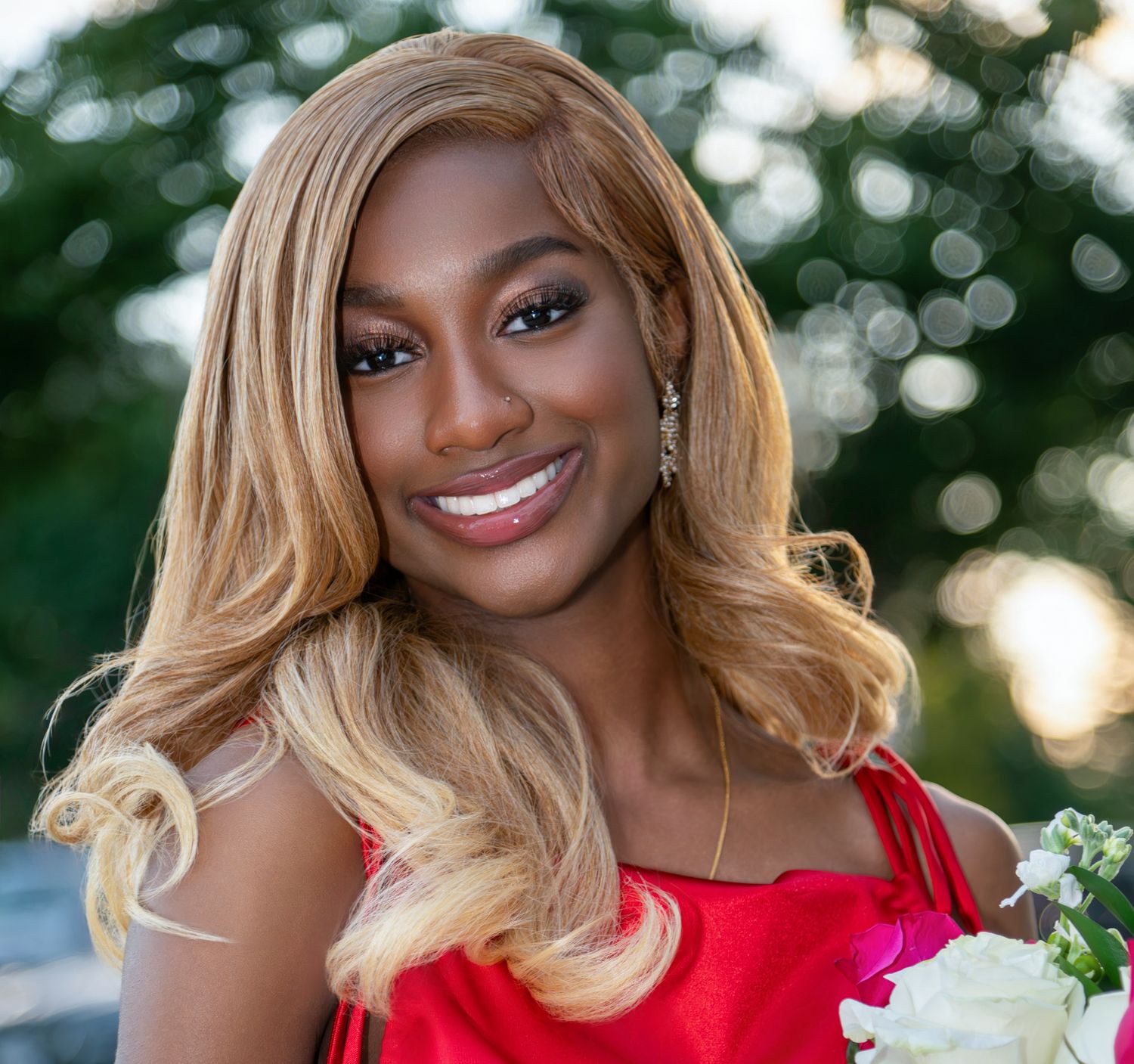 Young woman with long blonde hair in a red dress smiling while holding white flowers outdoors at golden hour.