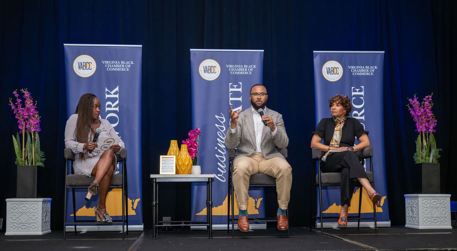 Three professionals engage in a panel discussion on stage at a business commerce networking event.
