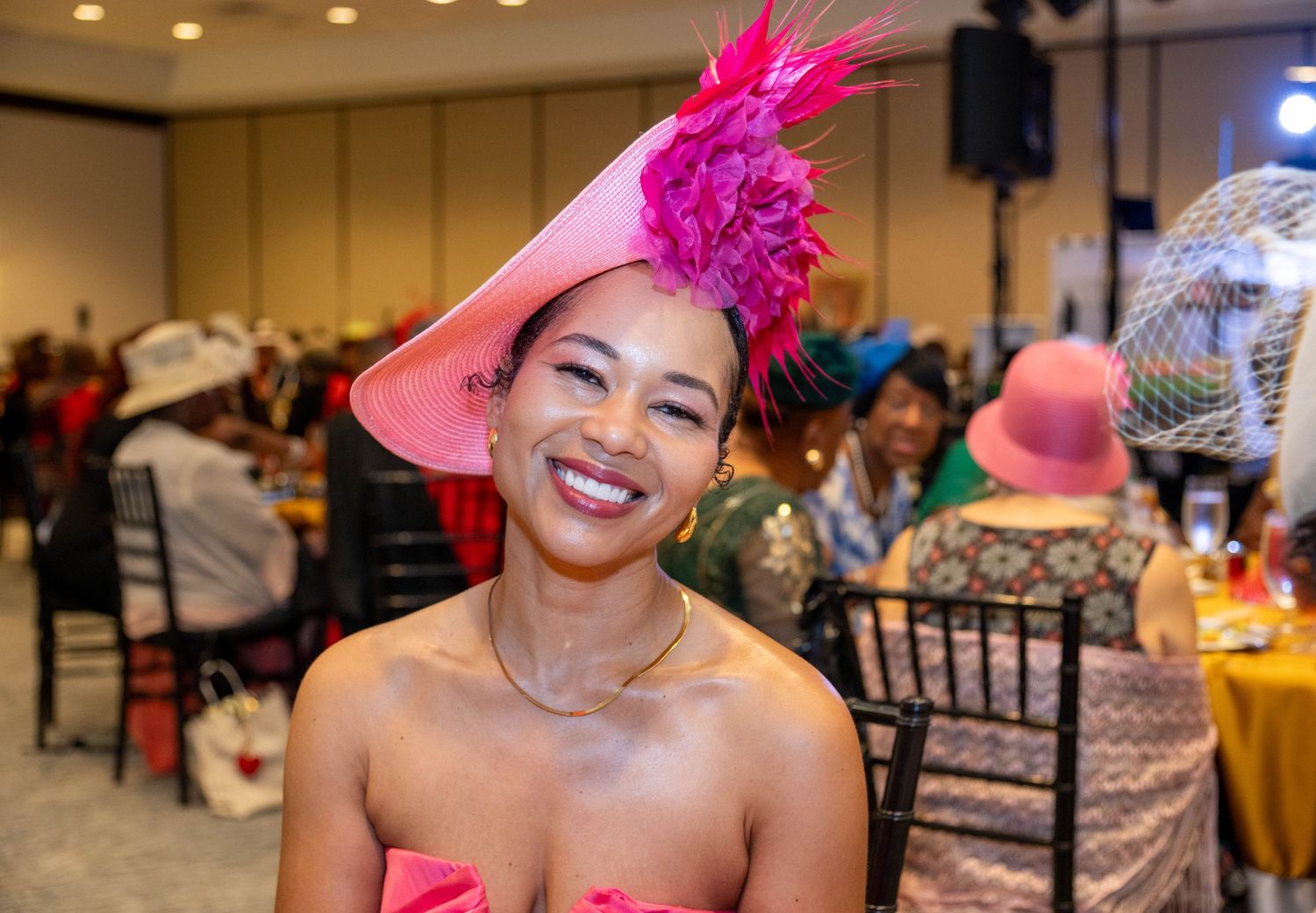 Woman smiling in pink strapless dress and floral feathered hat at elegant indoor event.