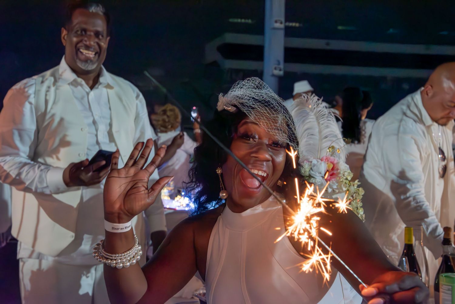 Woman in white dress holding sparkler at festive party celebration with guests dressed in white attire.
