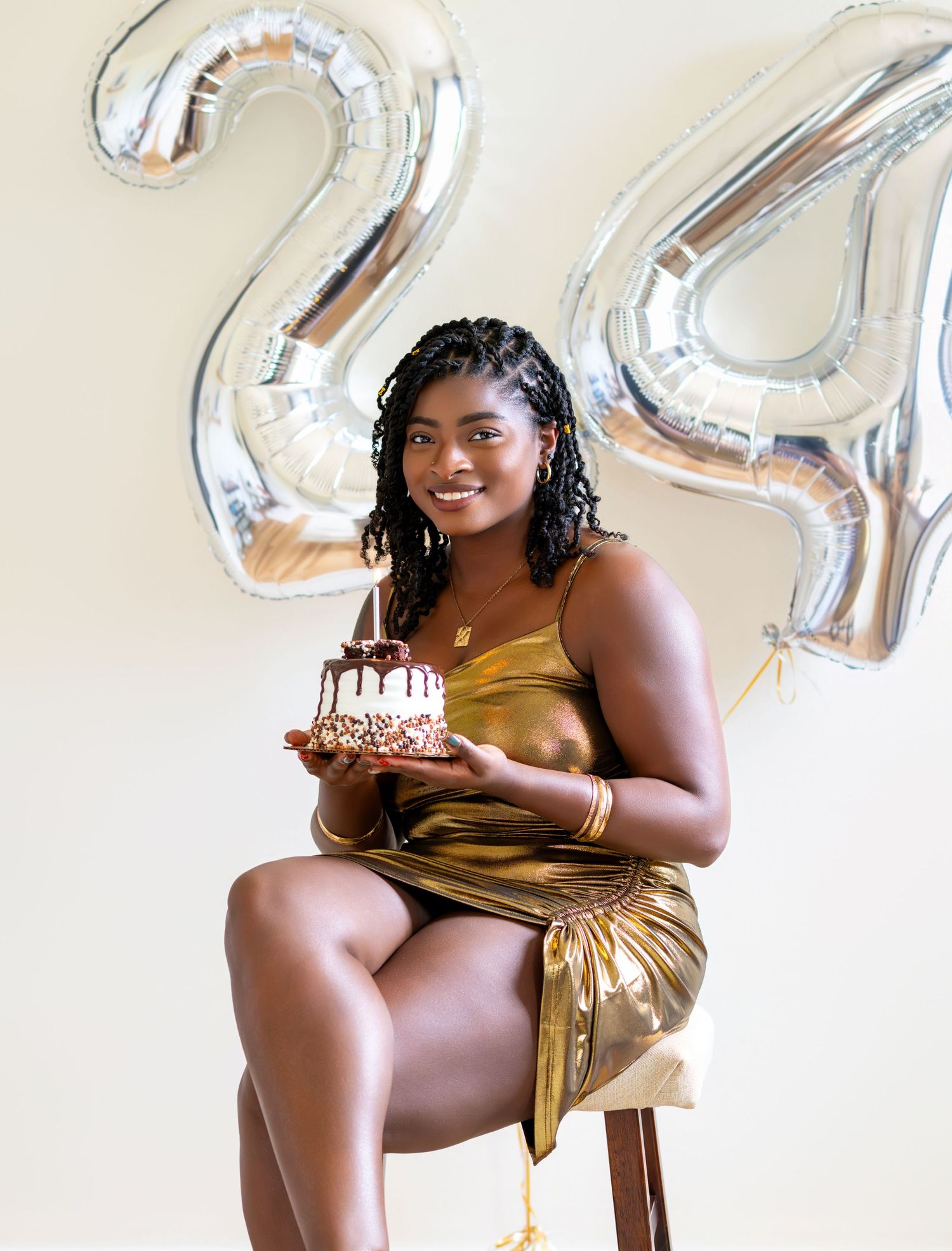 Woman in gold dress holding birthday cake in front of number 24 silver balloons.