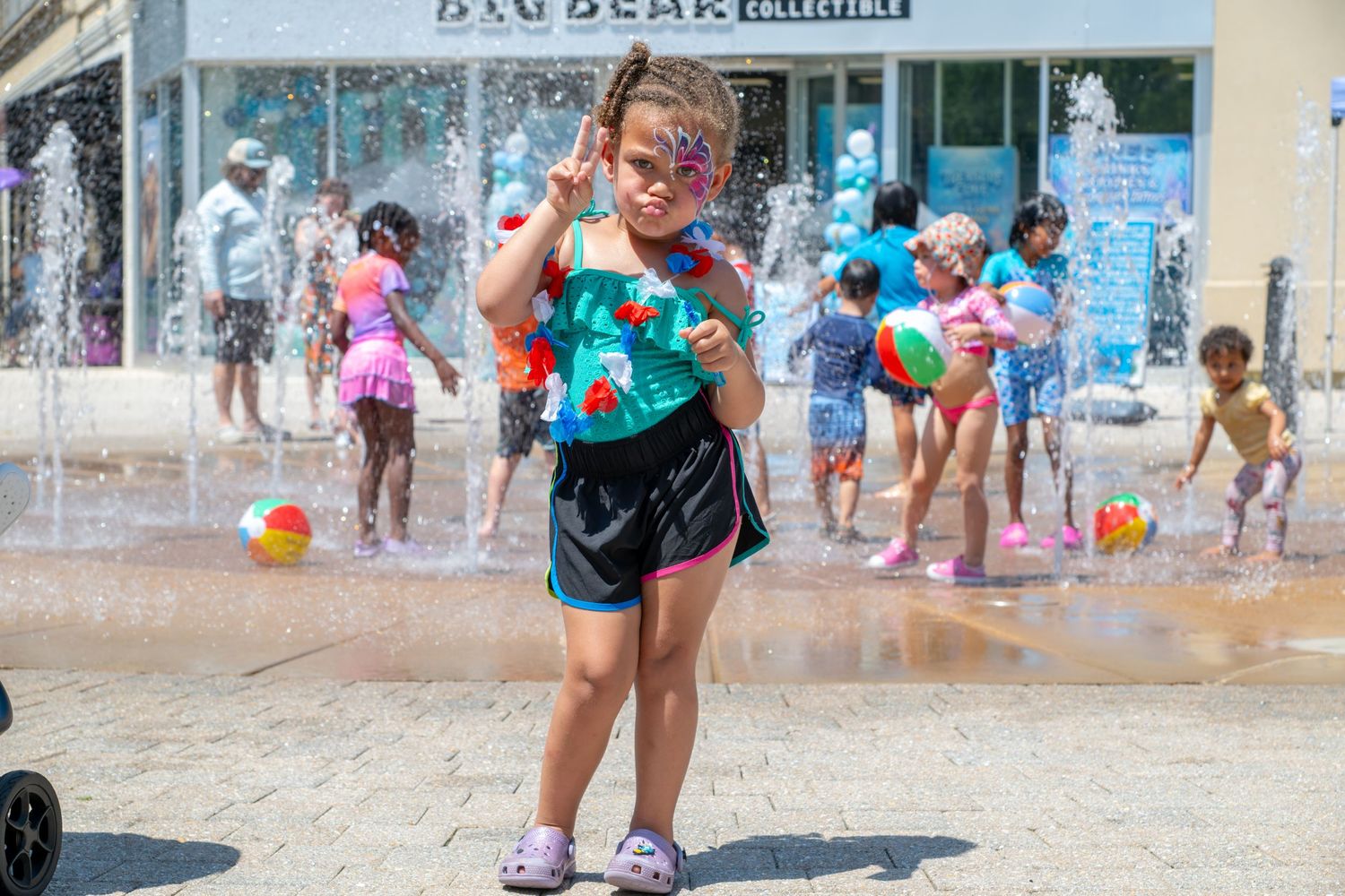 Young girl in teal swimsuit plays at outdoor splash pad fountain on sunny day with other children in background.