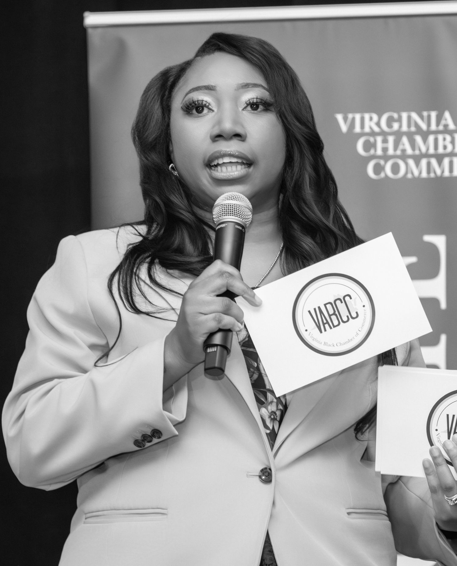 Woman speaking at Virginia Chamber of Commerce event, holding microphone and VABCC sign, black and white photo.