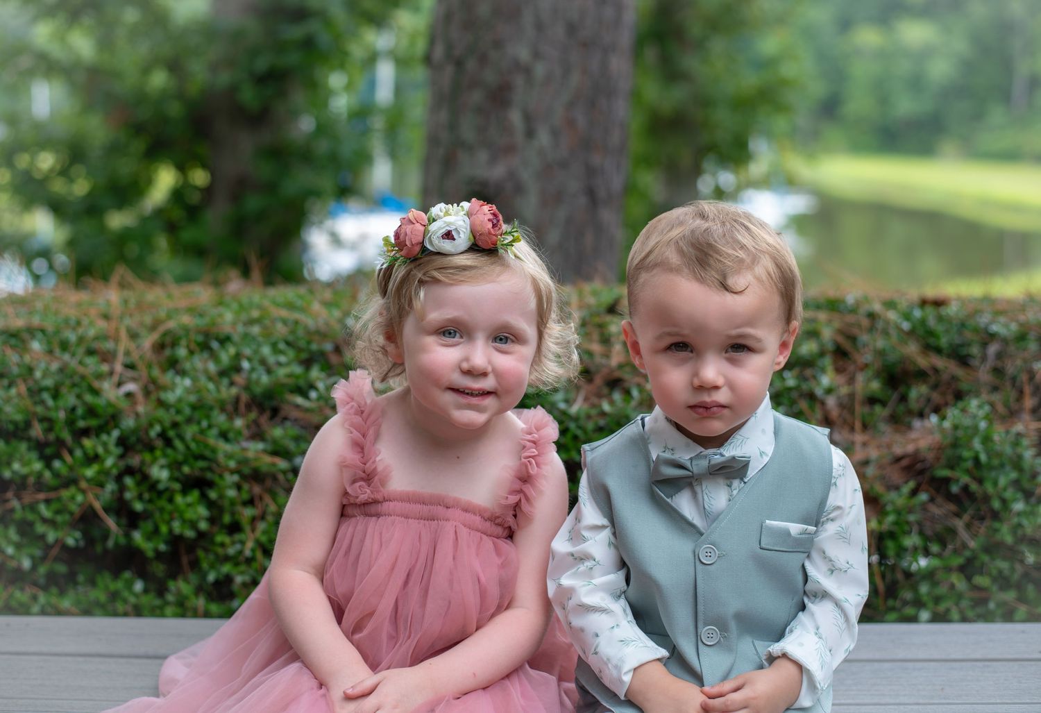 Two young children dressed formally sitting together, girl in pink dress with flower crown and boy in gray suit with bow tie.