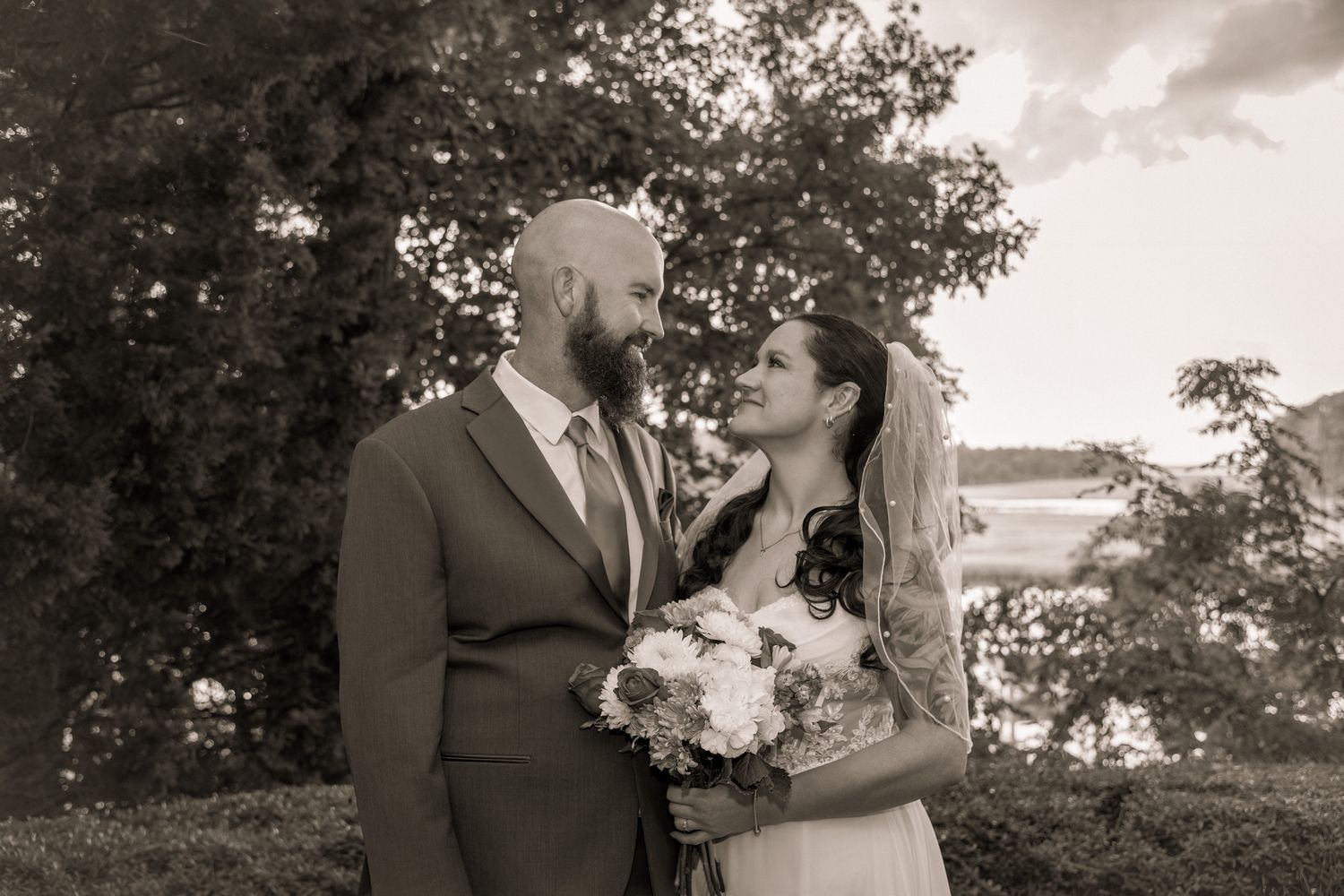 Black and white wedding photo of a bearded groom and veiled bride holding a bouquet, gazing at each other outdoors.