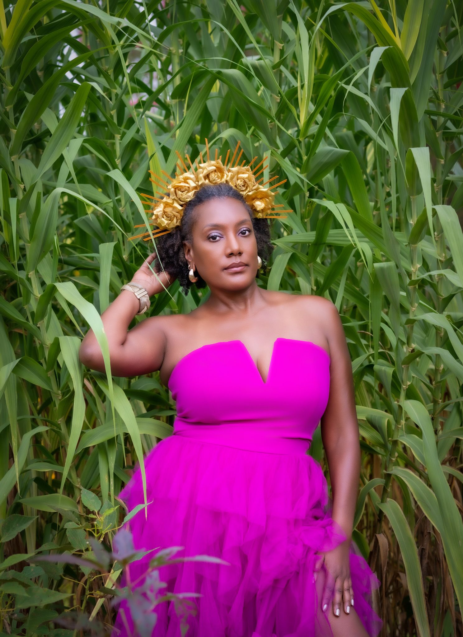 Woman in vibrant pink strapless gown wearing a gold floral crown standing among tall green corn stalks.