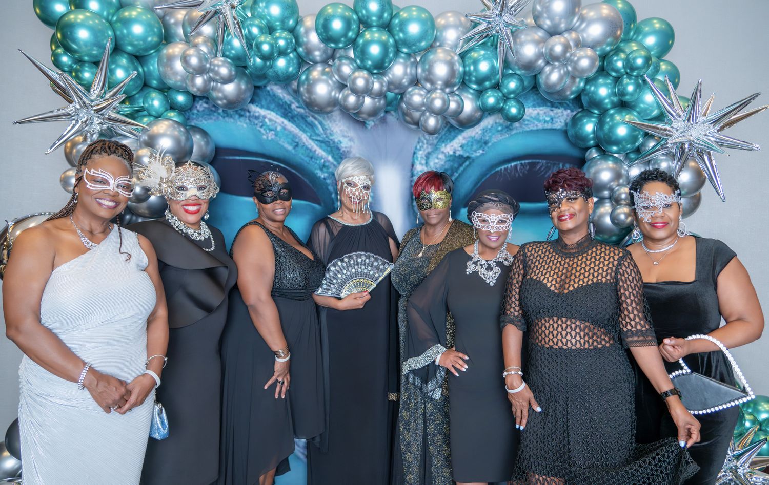 Group of women in black dresses and masquerade masks posing in front of teal balloon arch at formal event.