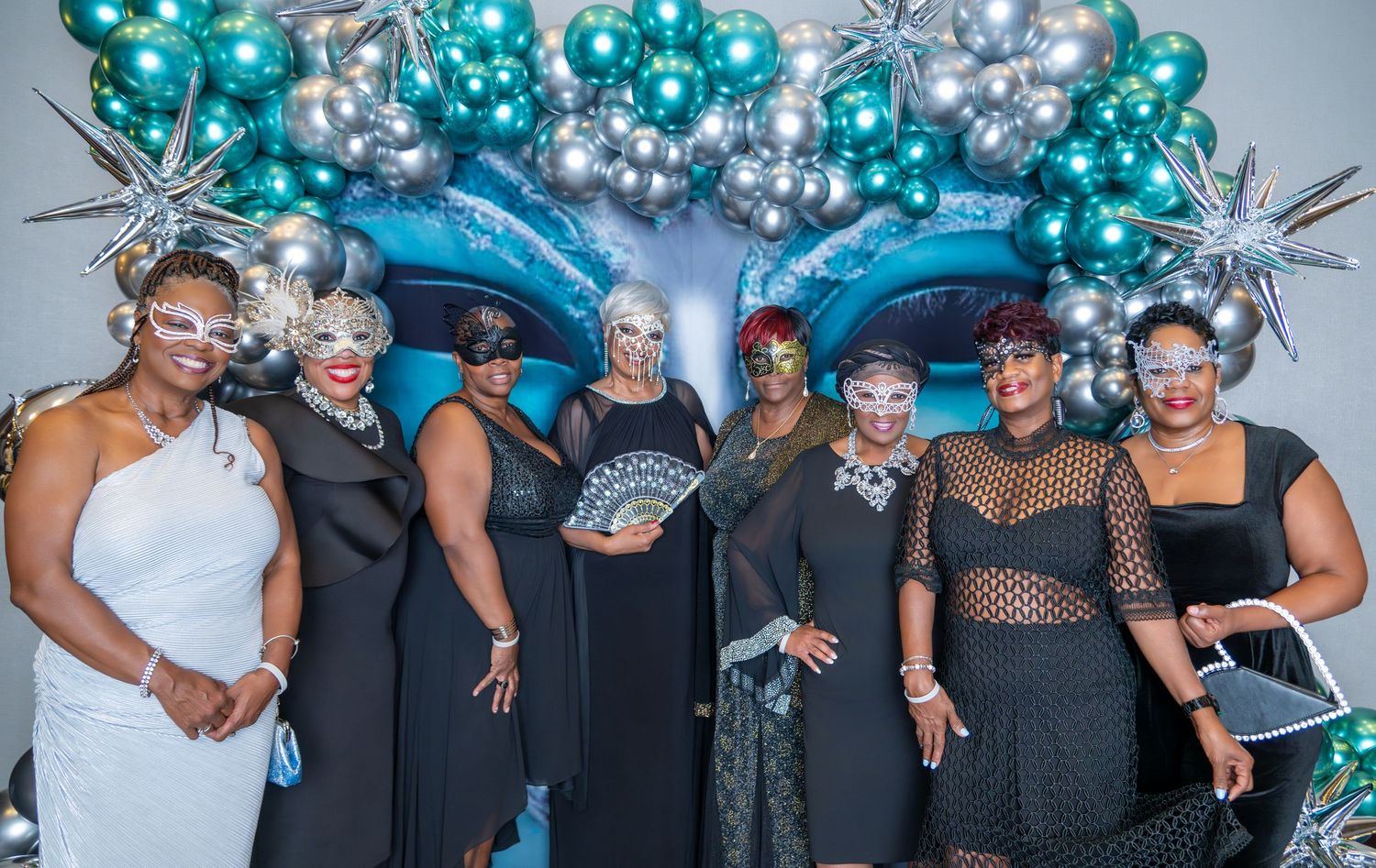 Group of women in elegant black outfits and masquerade masks posing at a teal balloon-decorated party event.