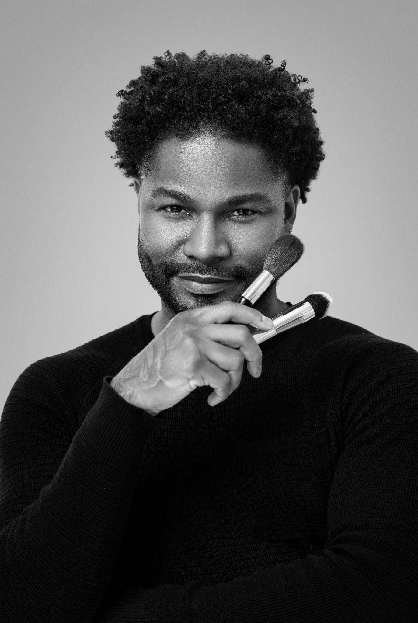 Black and white portrait of a man holding makeup brushes near his face wearing a black turtleneck sweater.