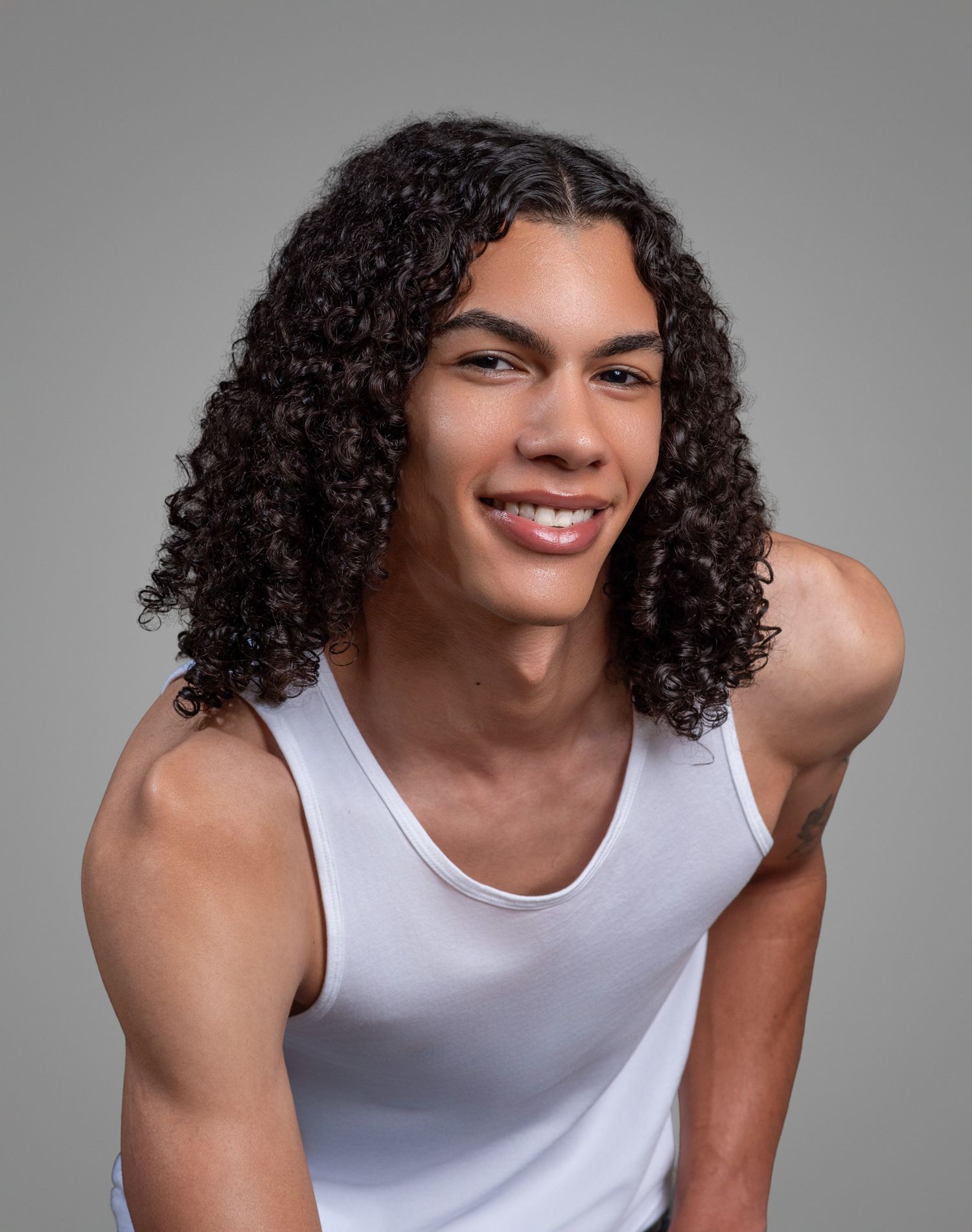 Smiling young man with long curly hair wearing a white tank top against a gray studio background.