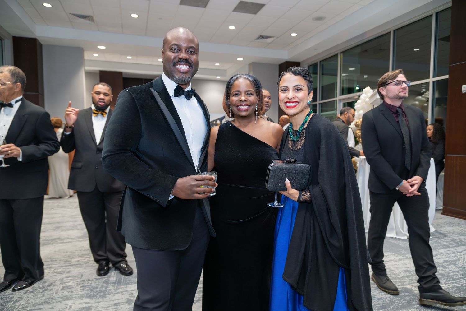 Three elegantly dressed guests smiling together at a formal gala event in a modern venue.