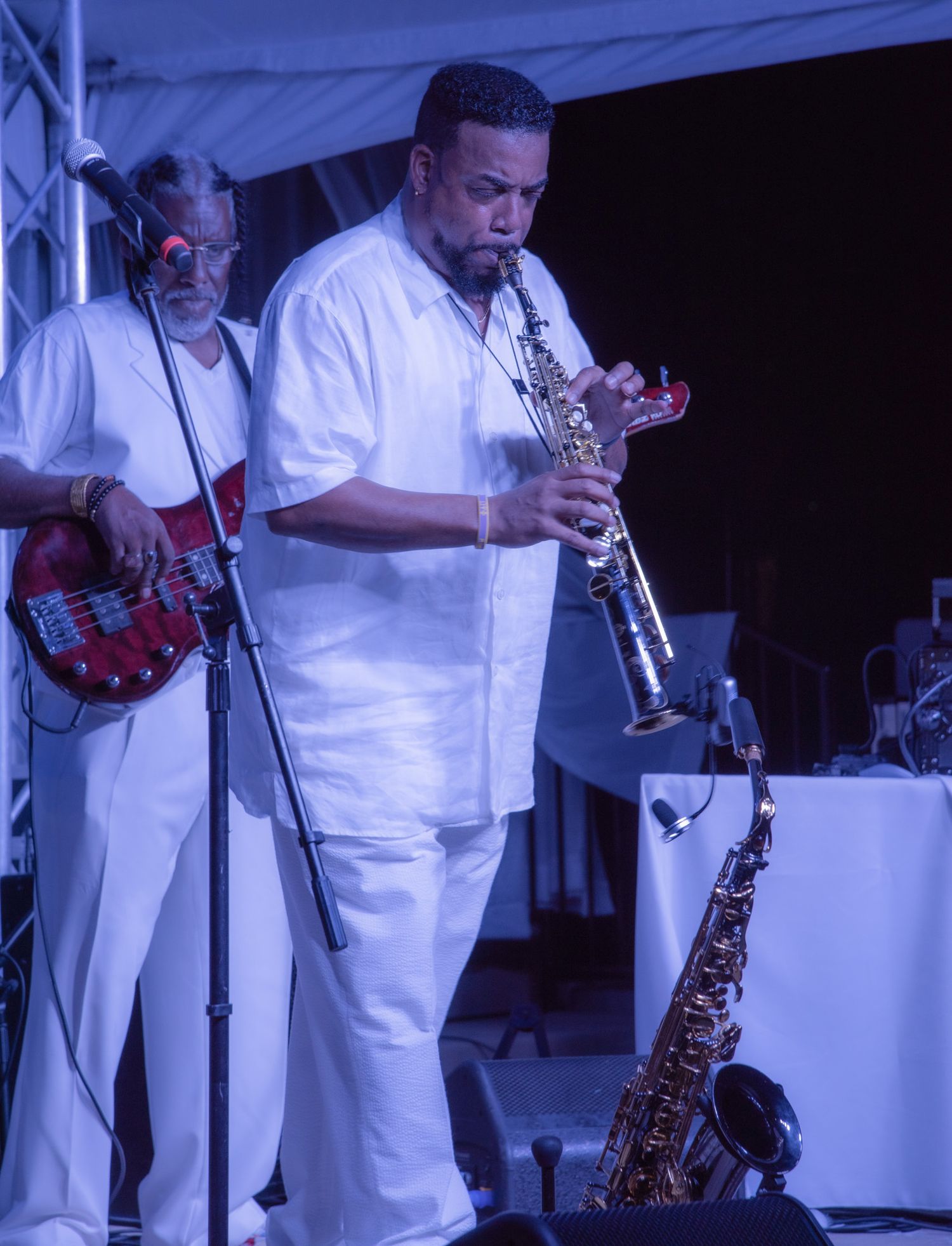 Musician in white outfit plays soprano saxophone on stage while bassist performs behind him at outdoor jazz concert.