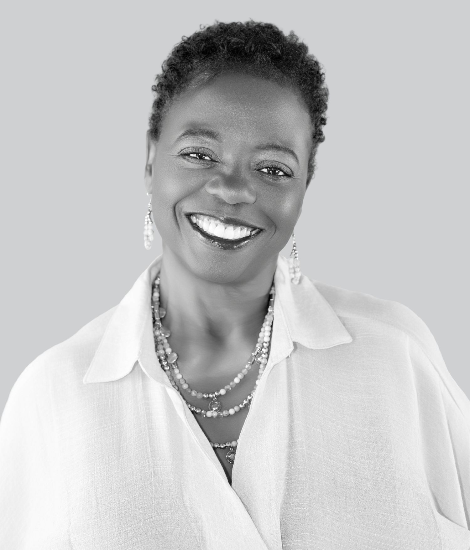Black and white portrait of a smiling woman with short hair wearing a white blouse and layered necklaces.