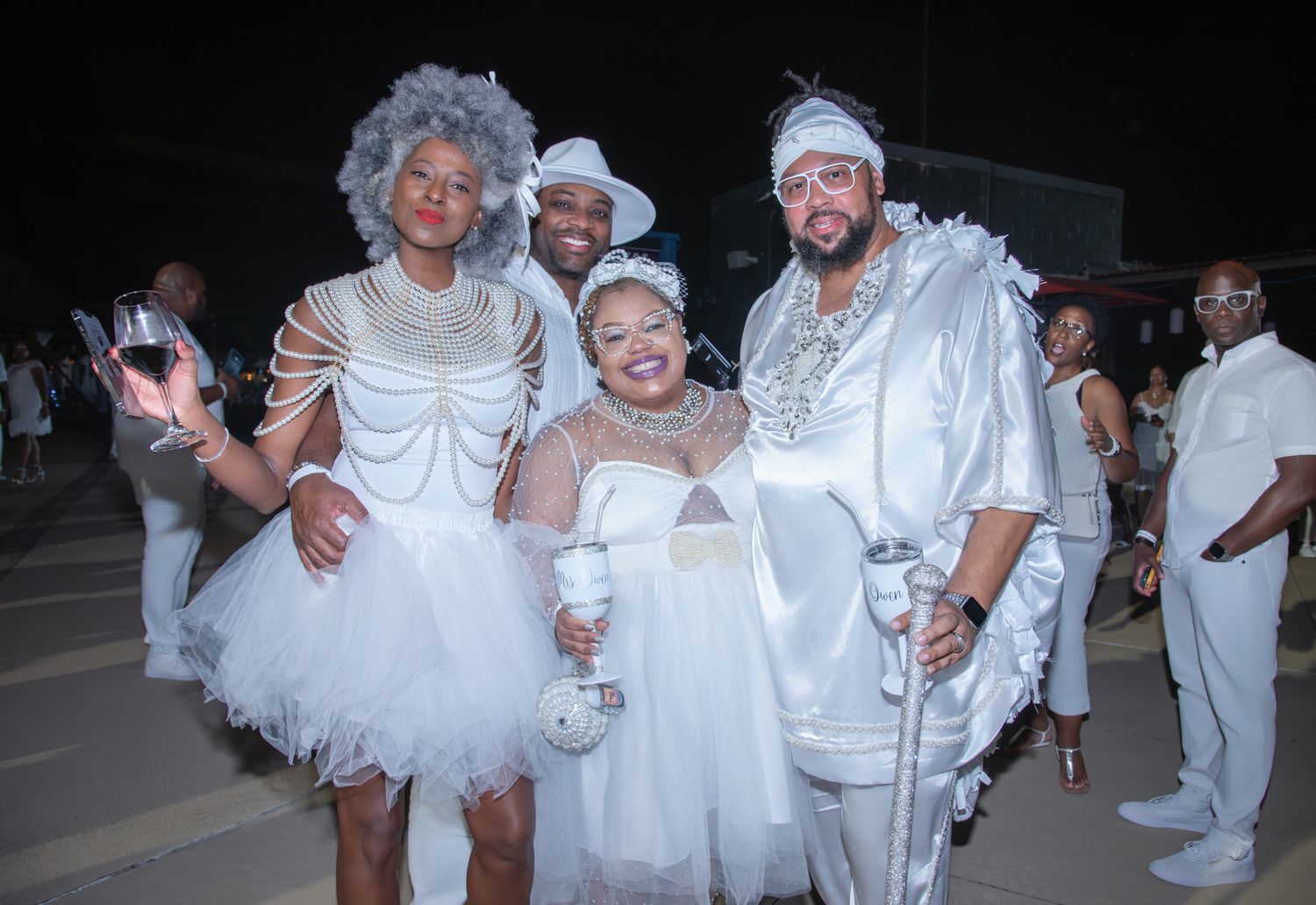 Four people in elegant white costumes and accessories pose together at a nighttime all-white themed party event.