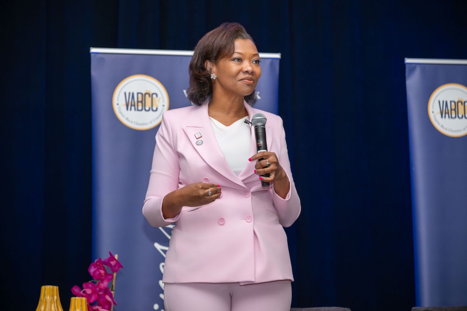 Woman in pink suit speaking at VABCC event, holding microphone on stage.