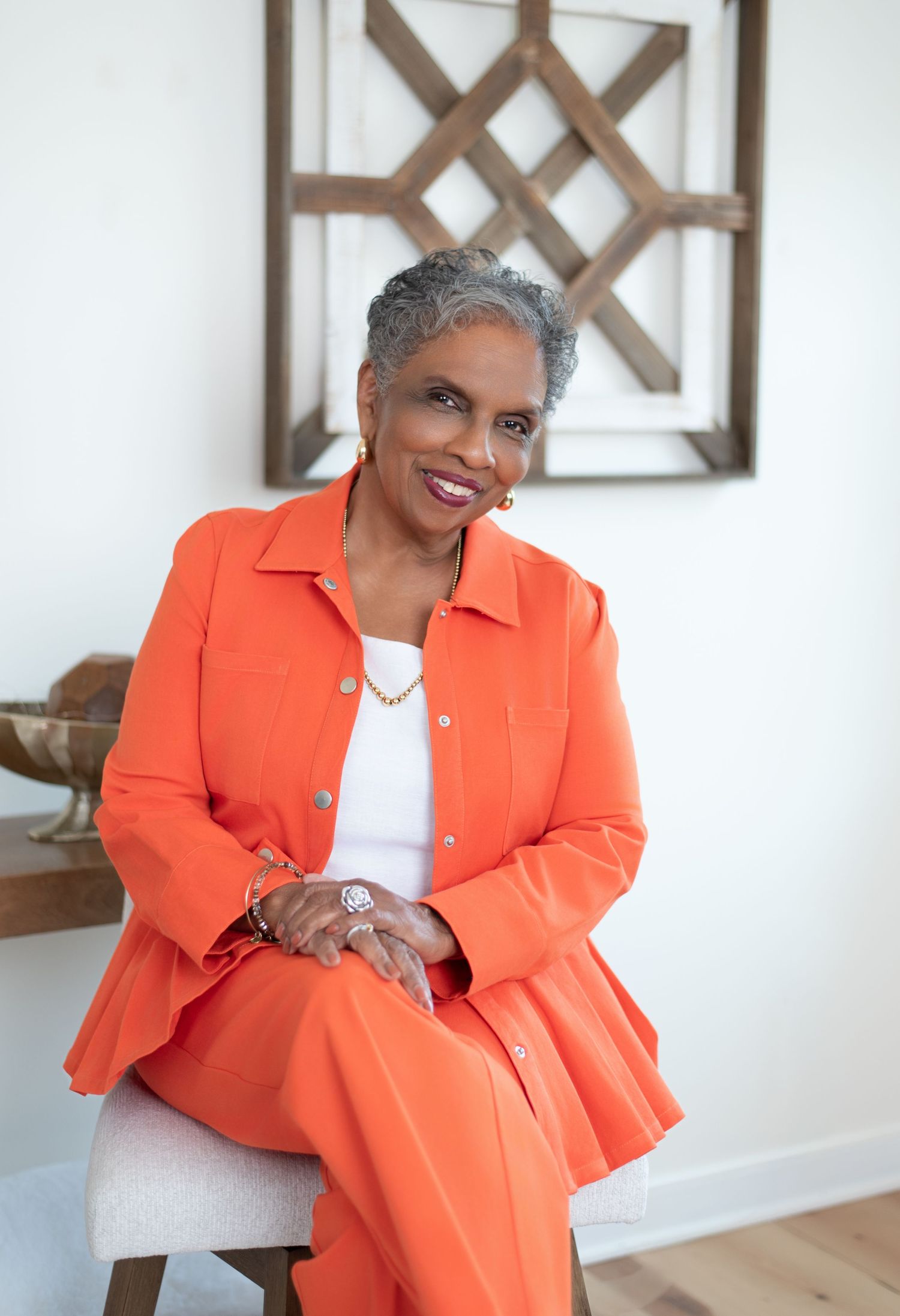 Stylish older woman in orange suit seated indoors smiling warmly near a decorative wall art piece.