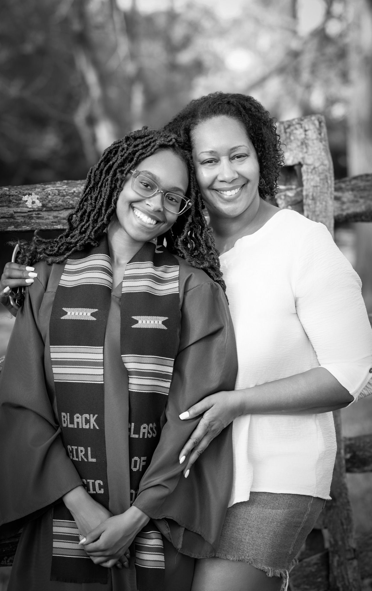 Two smiling women pose together outdoors, one wearing a graduation gown and kente cloth stole at a commencement celebration.