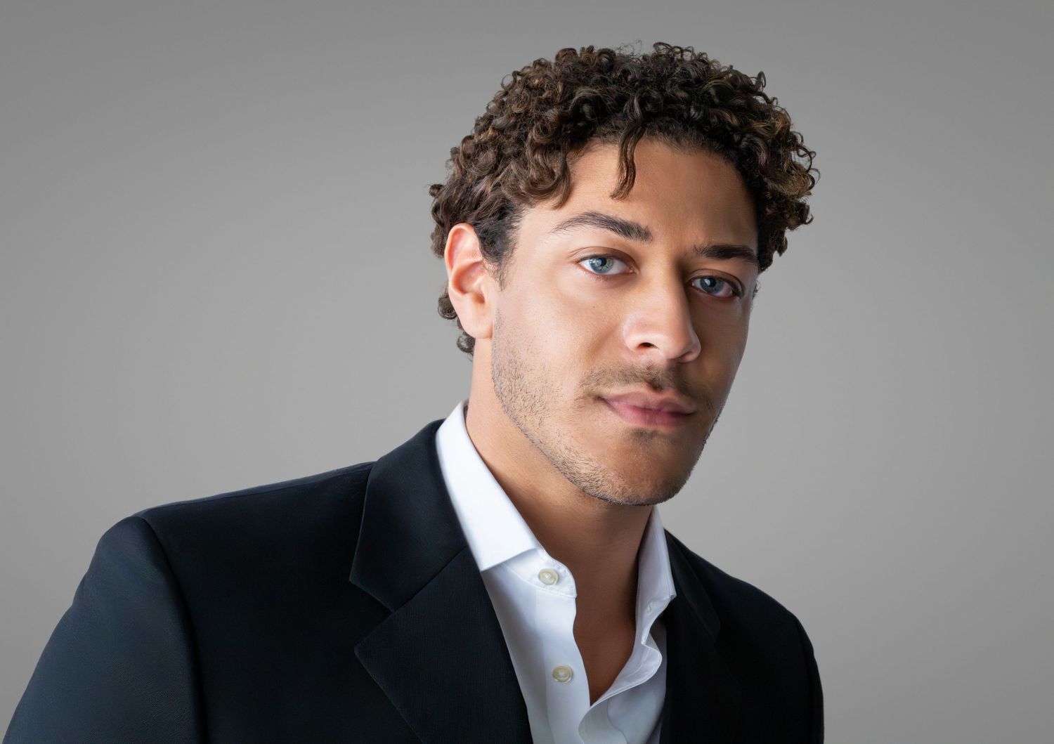 Young man with curly hair in a black blazer and white shirt posing against a gray studio background.