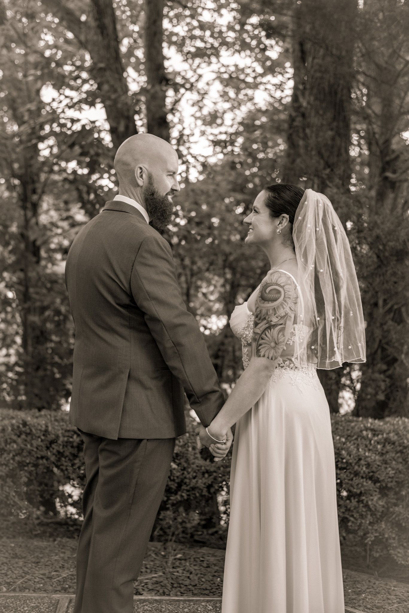 Black and white photo of a bride and groom holding hands outdoors among trees on their wedding day.