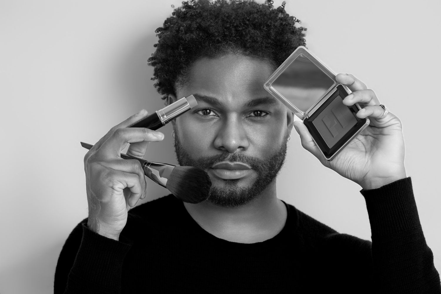 Black and white portrait of a man holding makeup brush and compact mirror against white background.