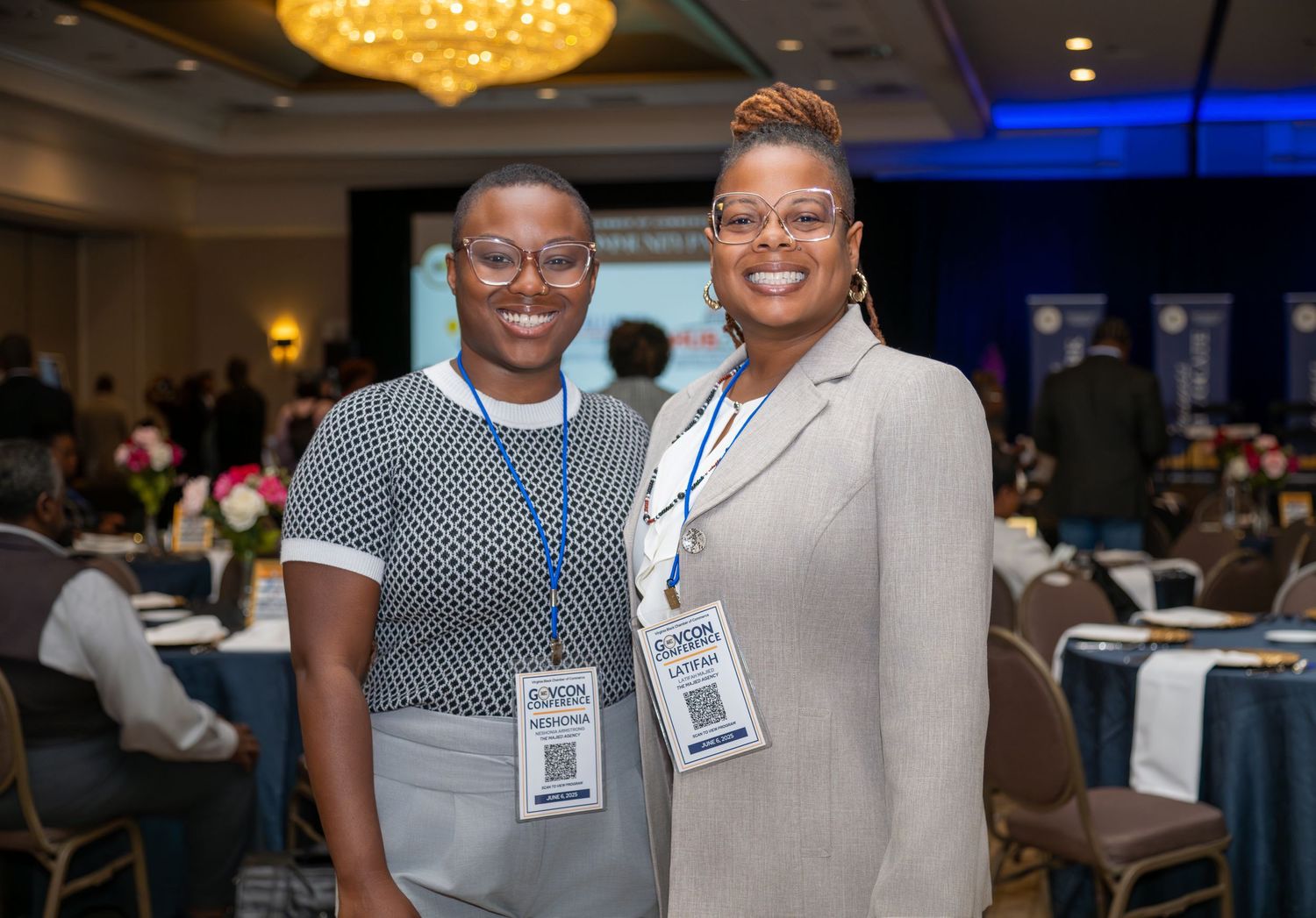Two professional women wearing lanyards smile together at a formal conference event in a ballroom.