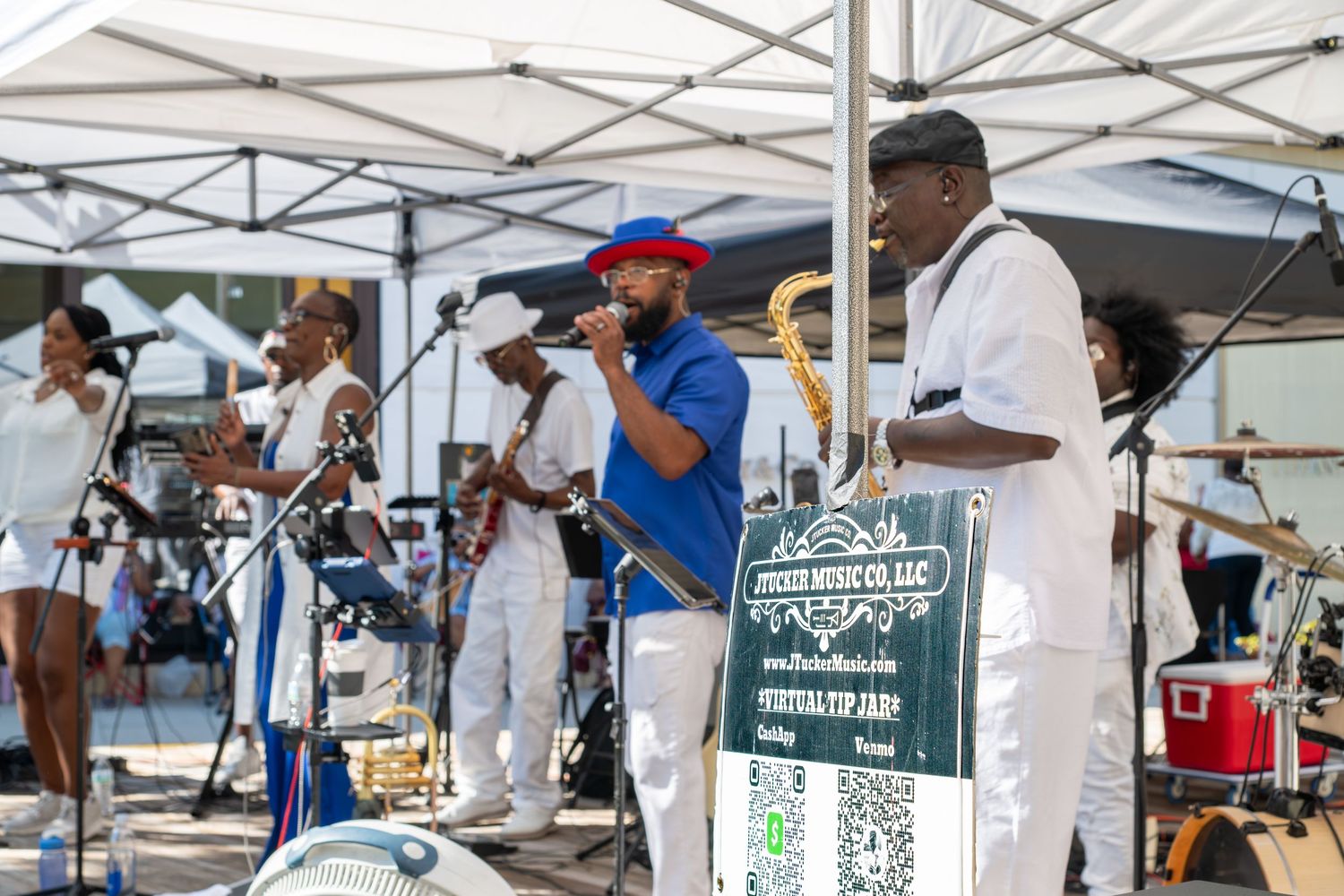 Live band performing outdoors at a festival, musicians in white outfits playing brass instruments under a tent.