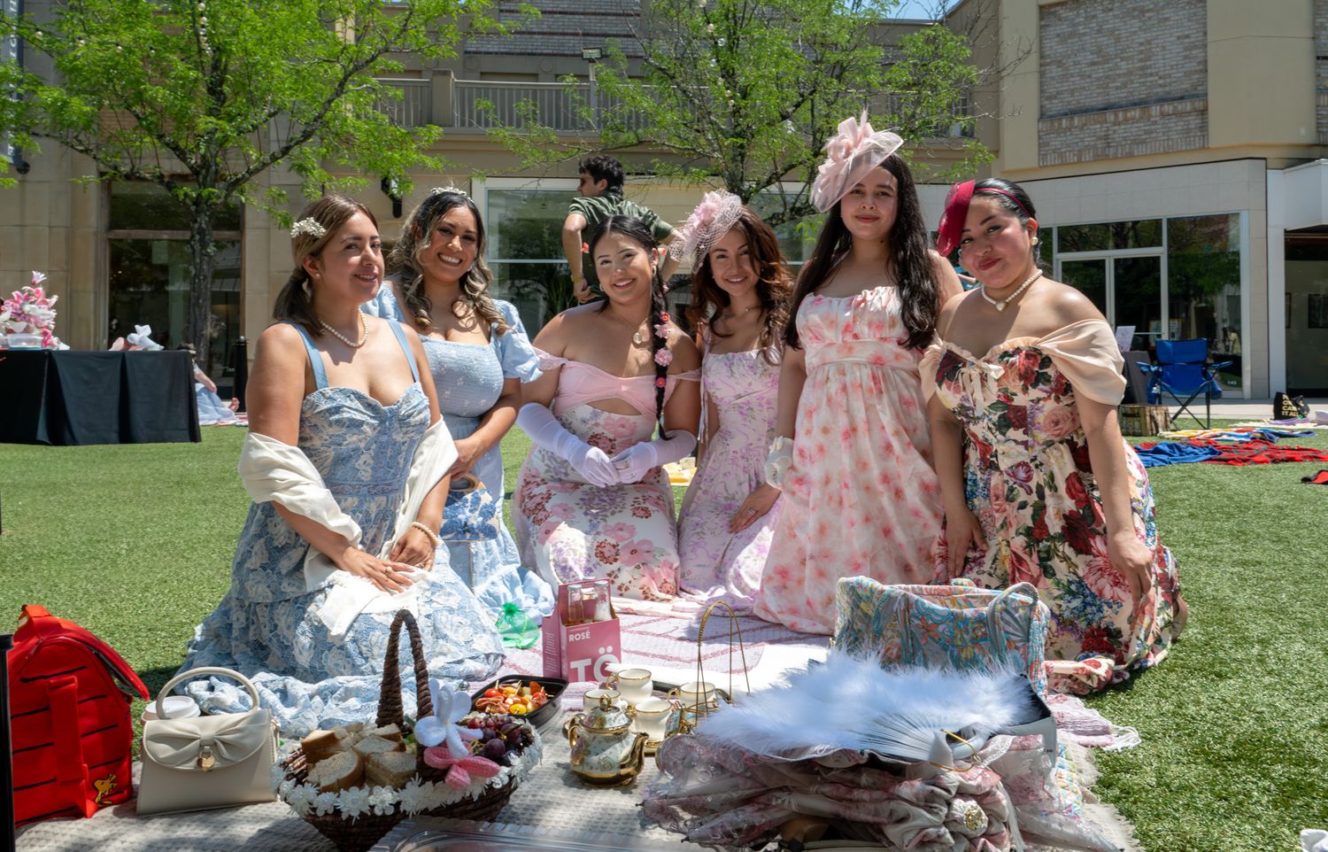 Six women in elegant floral dresses pose together outdoors at a garden picnic party on a sunny day.