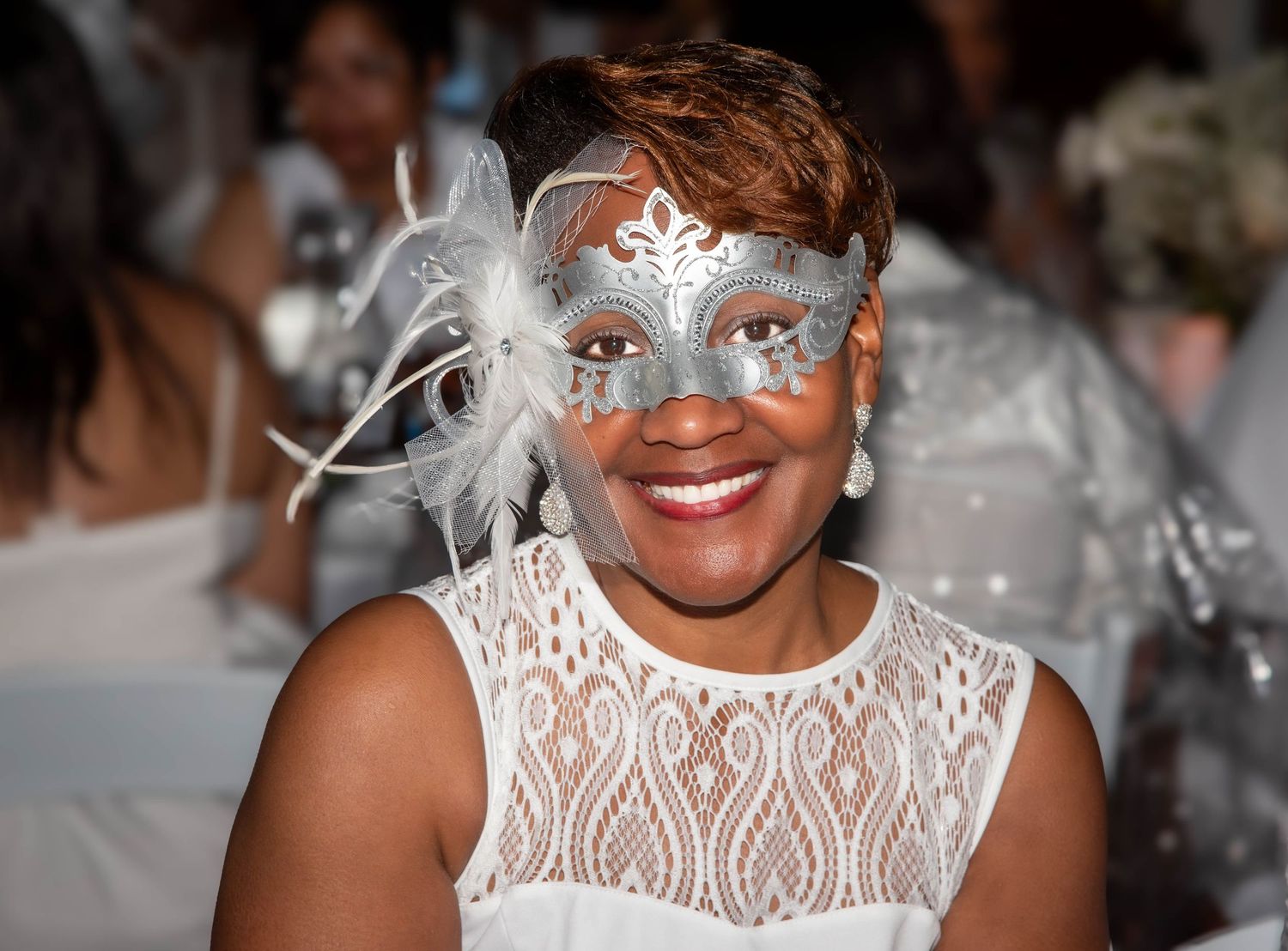 Woman smiling in silver masquerade mask with feathers at formal white party event.