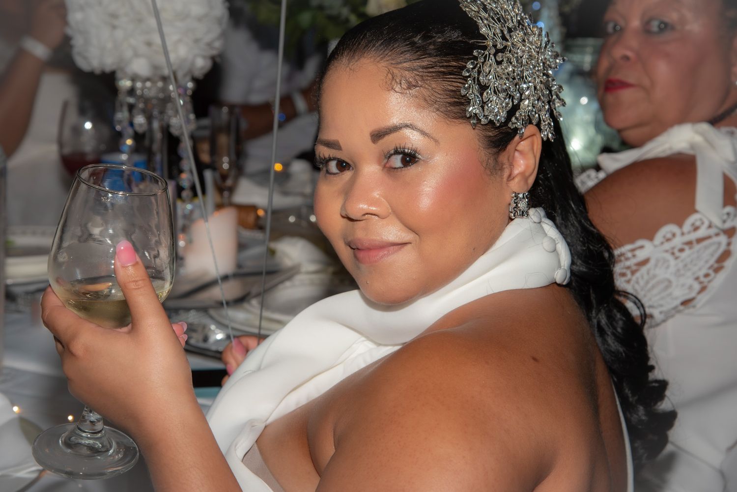 Woman in white dress holding wine glass at elegant event, wearing decorative hair accessory.
