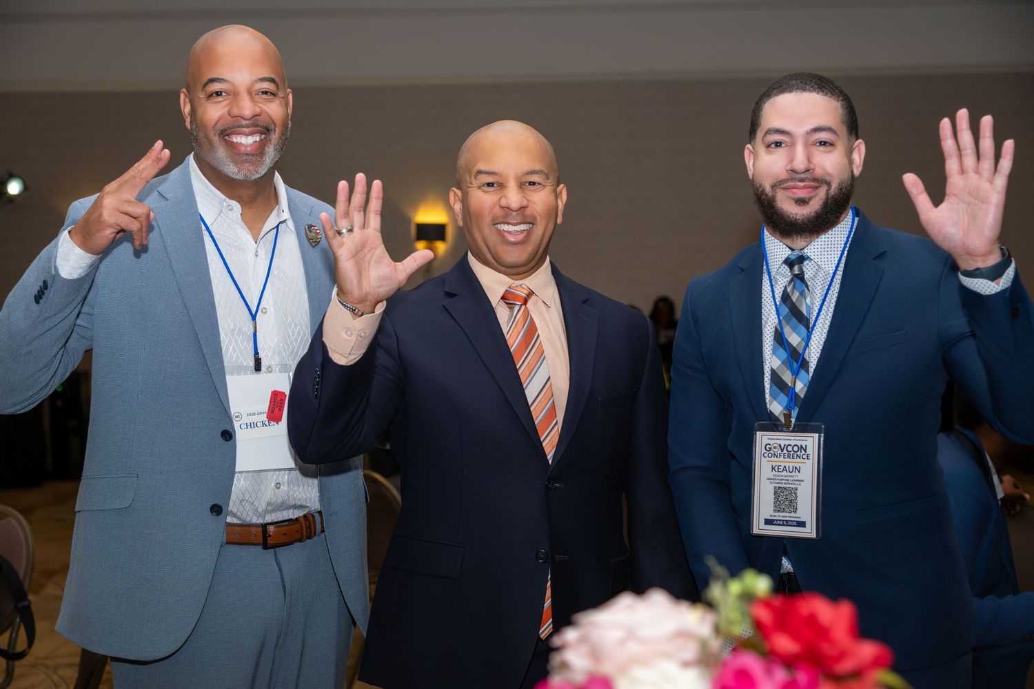 Three men in suits smiling and waving at a formal event or conference, wearing name badges.
