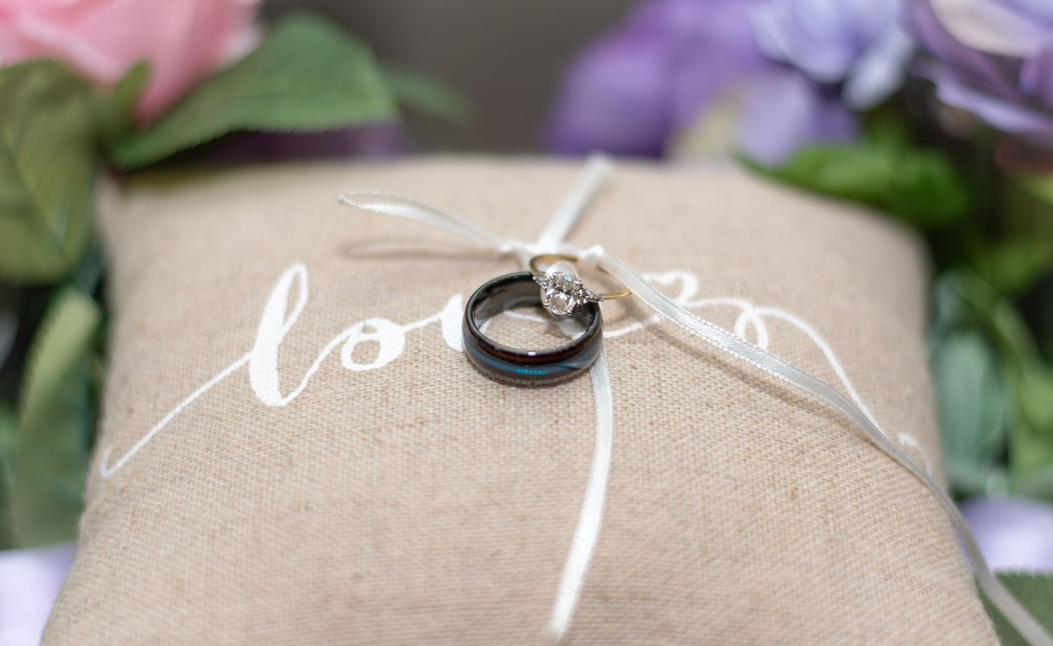 Wedding rings on a burlap pillow with white ribbon and calligraphy script surrounded by purple and green floral decor.