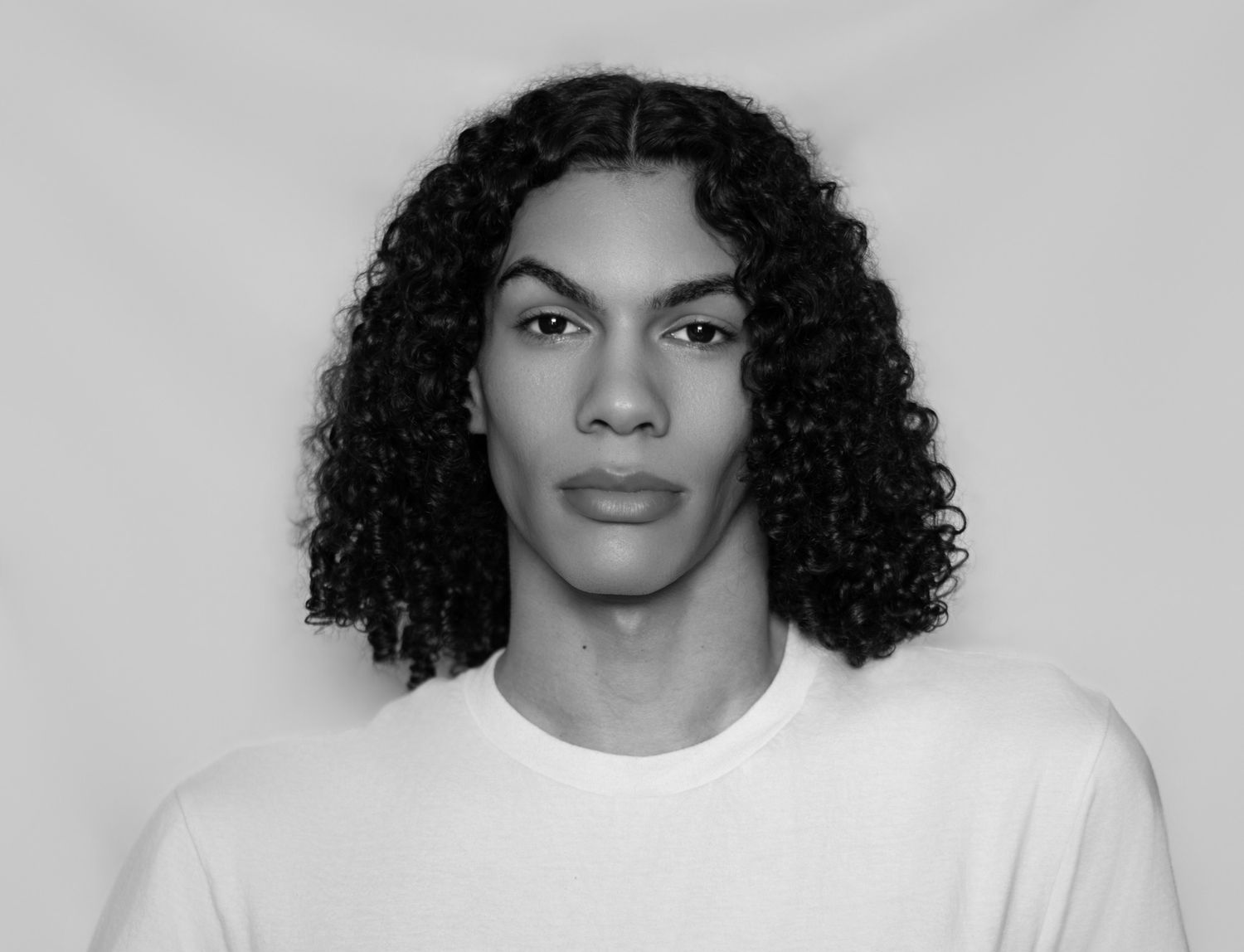 Black and white portrait of a young man with long curly hair wearing a white t-shirt against a light background.